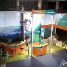 trk system stoiska targow i handlowe - Widok z góry na stoisko konferencyjne GSK z logo Flixoide i Serevent, wyposażone w dwa pomarańczowe lady z czarnymi hokerami, oświetlenie punktowe i dekoracyjne rośliny w doniczkach.