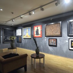 trk system stoiska targow i handlowe - Wnętrze galerii sztuki z szarą ścianą, na której wiszą różnorodne obrazy w ramach, oświetlone reflektorami, z drewnianą podłogą, sofą i stolikami kawowymi z wazonami.