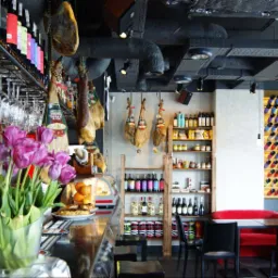 Wnętrze baru tapas z szynkami wiszącymi nad barem, kwiatami w wazonie, półkami z produktami i dekoracyjną ścianą z butelkami wina.