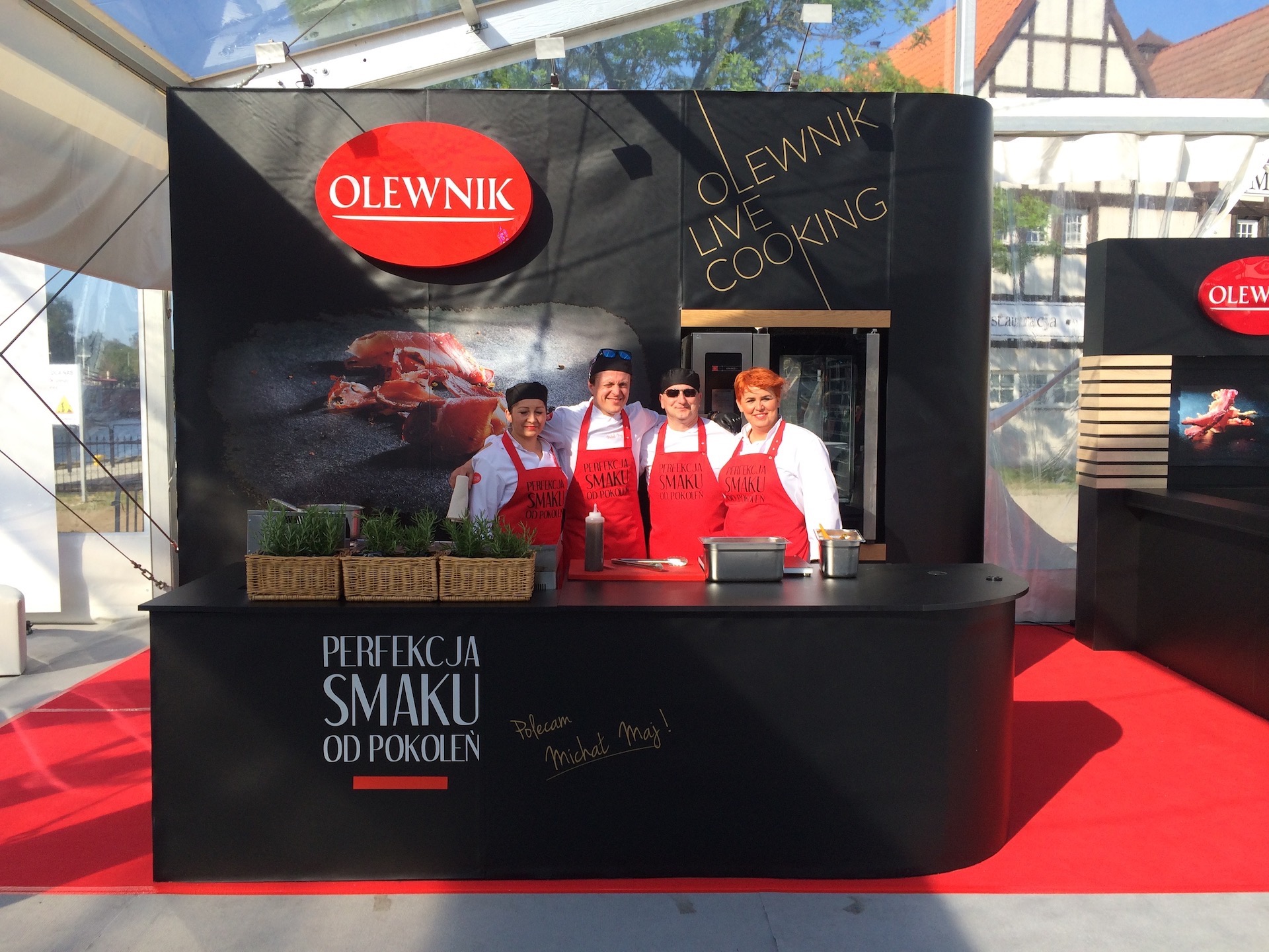 Stoisko reklamowe Olewnik z warsztatami kulinarnymi live cooking, prezentujące zespół kucharzy w czerwonych fartuchach na tle czarnego panelu z logo i hasłem 'Perfekcja smaku od pokoleń'.