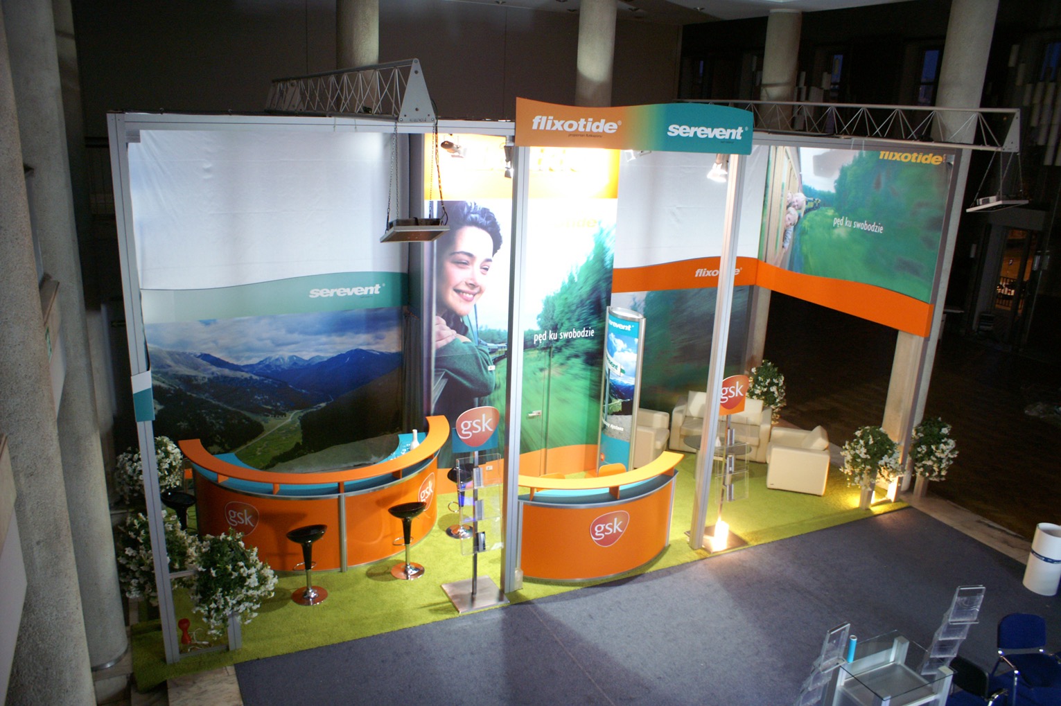 Widok z góry na stoisko konferencyjne GSK z logo Flixoide i Serevent, wyposażone w dwa pomarańczowe lady z czarnymi hokerami, oświetlenie punktowe i dekoracyjne rośliny w doniczkach.