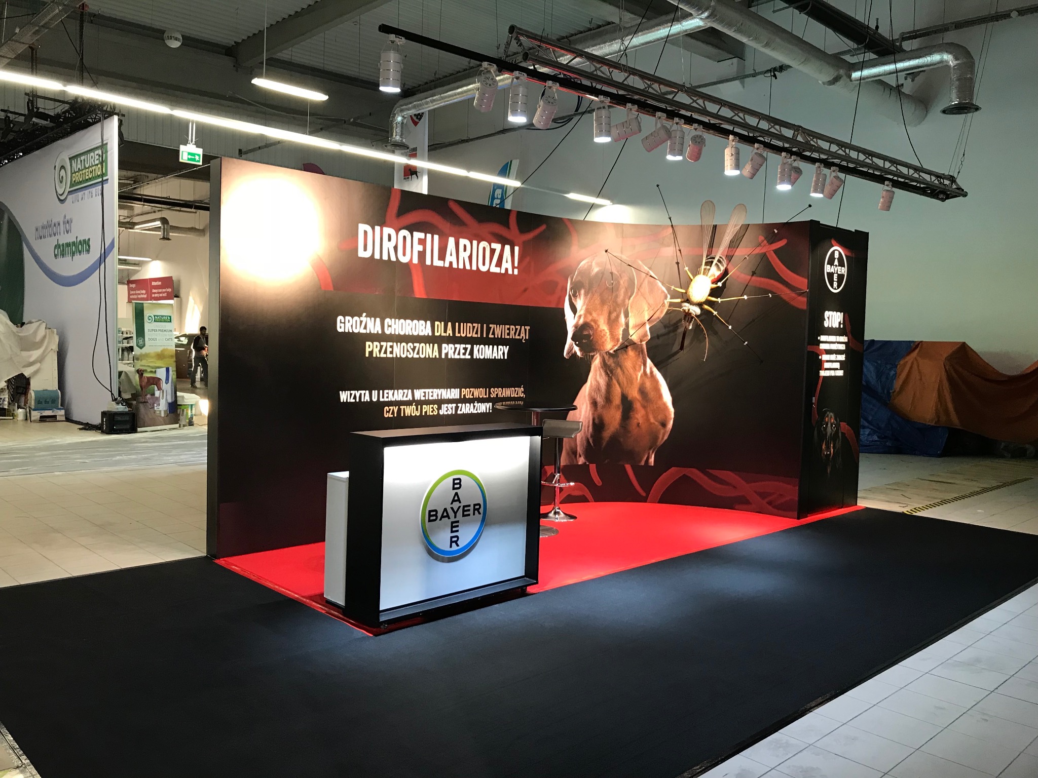 Stoisko konferencyjne Bayer z grafiką przedstawiającą psa i komara, informujące o dirofilariozie, oświetlone reflektorami na tle surowego wnętrza hali.