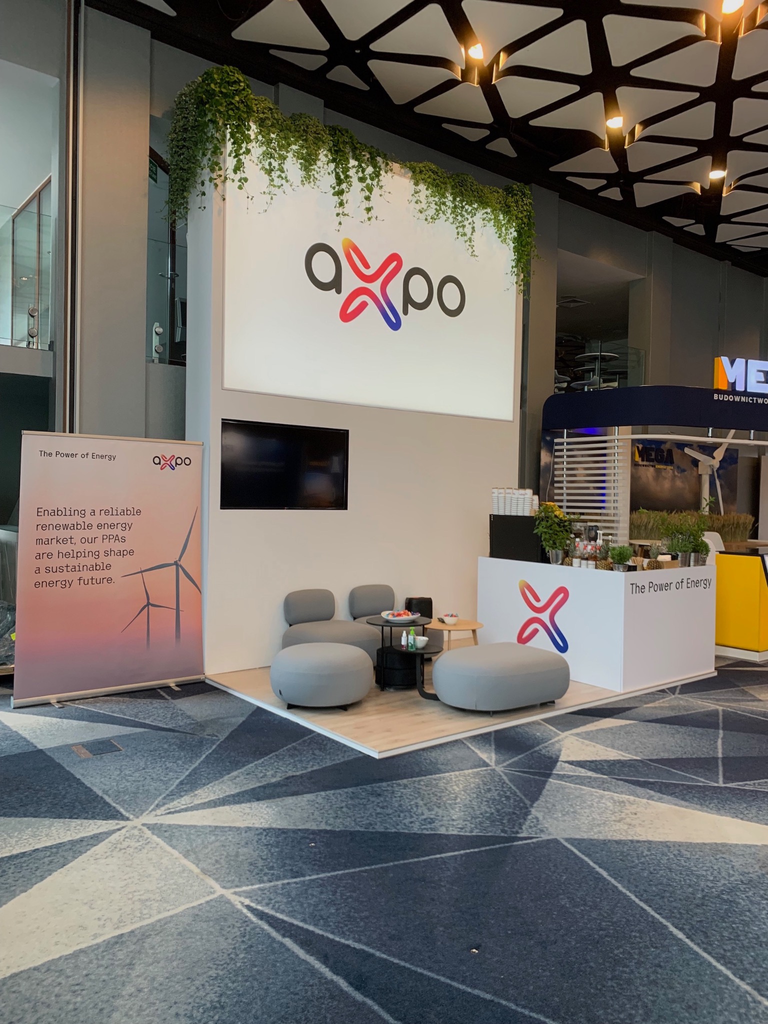 Stoisko konferencyjne Axpo w Hotelu Narvil, z logo firmy, roślinami zwisającymi z góry, ekranem i meblami wypoczynkowymi, na tle wzorzystej wykładziny.