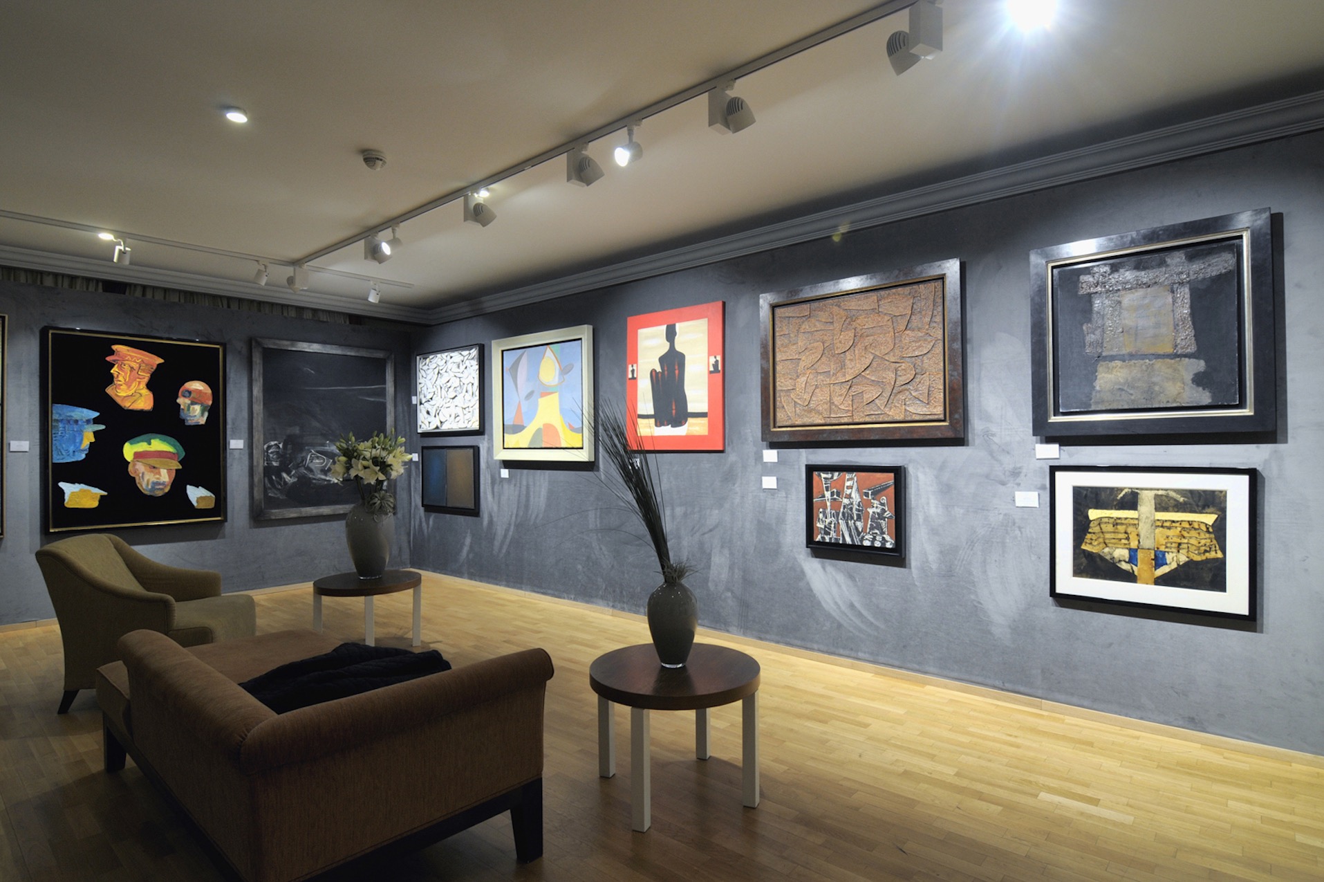Wnętrze galerii sztuki z szarą ścianą, na której wiszą różnorodne obrazy w ramach, oświetlone reflektorami, z drewnianą podłogą, sofą i stolikami kawowymi z wazonami.