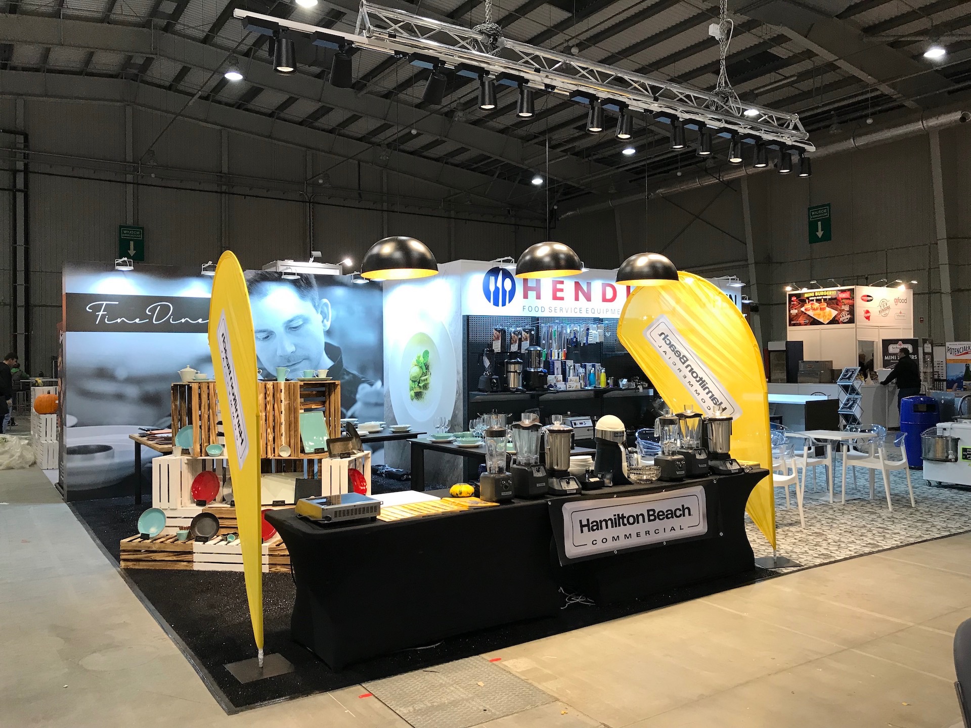 Stoisko targowe prezentujące sprzęt gastronomiczny marek Hendi i Fine Dine, z ekspozycją blenderów Hamilton Beach Commercial, oświetlone reflektorami podwieszonymi pod sufitem hali.