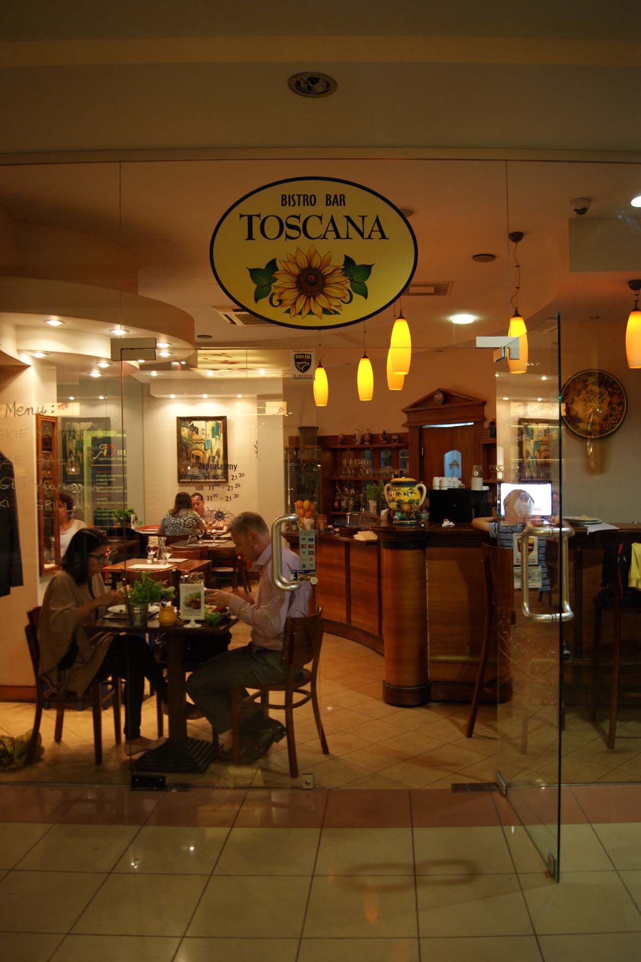 Wnętrze Bistro Bar Toscana z drewnianym barem, wiszącymi lampami i klientami przy stolikach, widoczne przez przeszklone wejście z logo firmy.
