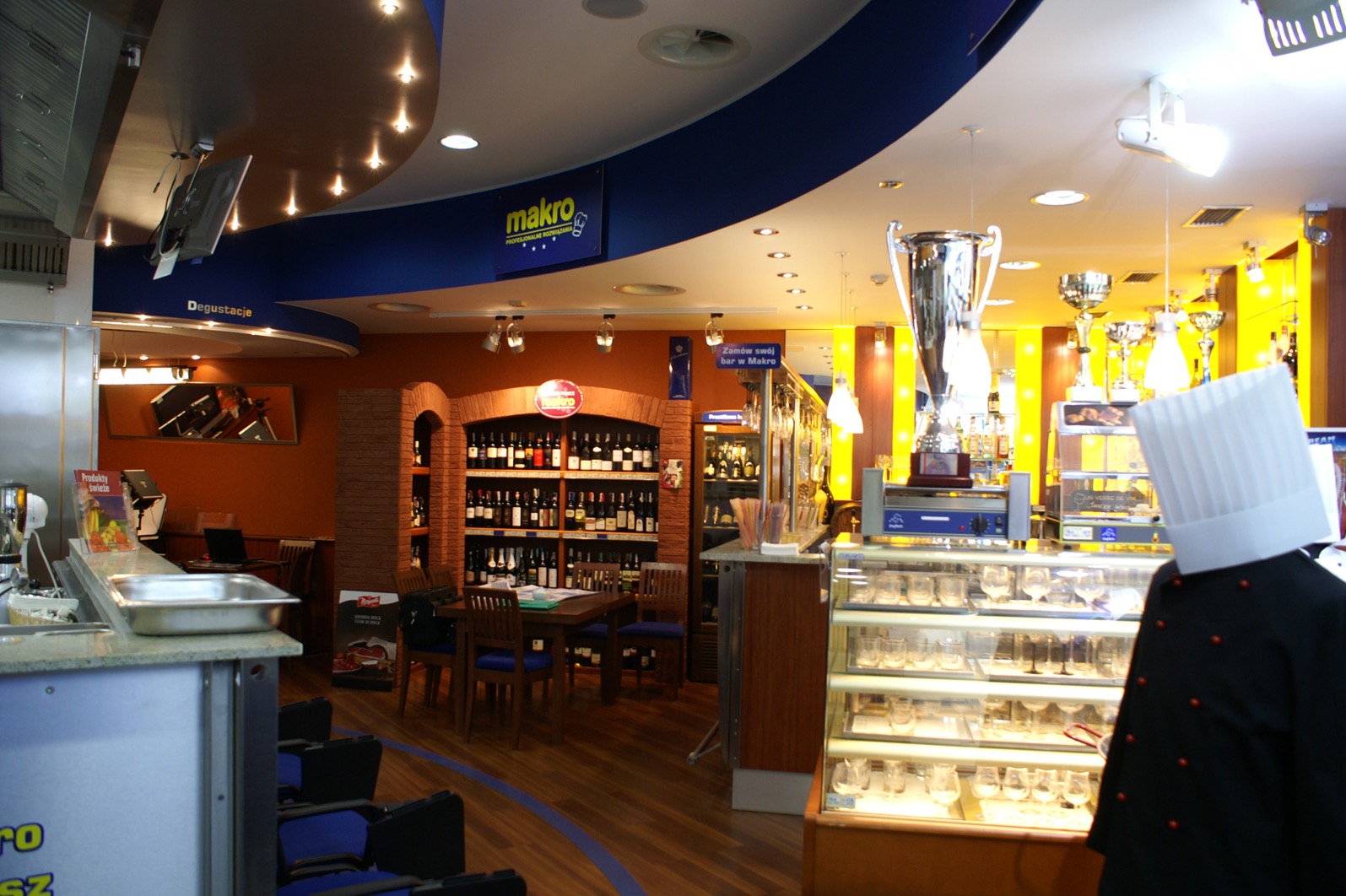 Wnętrze restauracji Makro Centrum HoReCa z ekspozycją win w regale z cegły, stołem z krzesłami oraz witryną chłodniczą na szkło, widoczny także kucharz z boku w uniformie i wysokiej czapce...