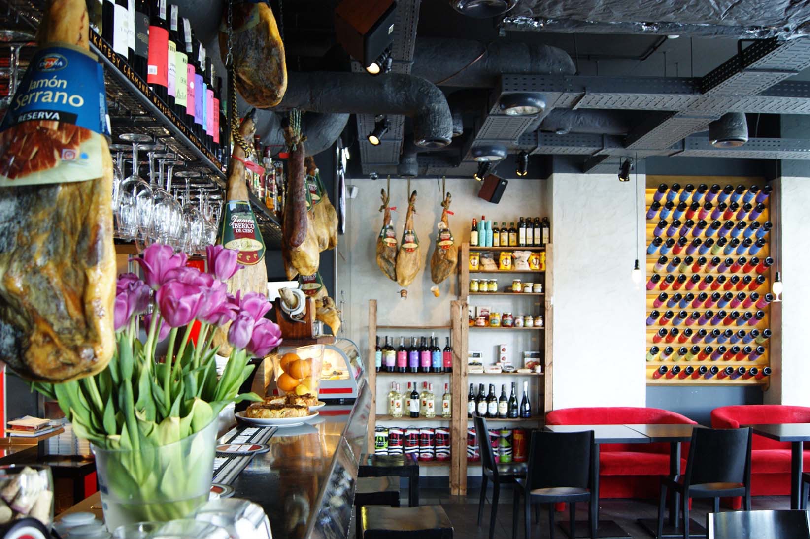 Wnętrze baru tapas z szynkami wiszącymi nad barem, kwiatami w wazonie, półkami z produktami i dekoracyjną ścianą z butelkami wina.