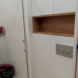 Remonty i wykończenia Kraków - Nowoczesna toaleta z białymi płytkami, zabudową meblową z drewnianą wnęką i chromowanym przyciskiem spłukiwania. W tle widoczna szklana kabina prysznicowa z wiadrem i mopem.