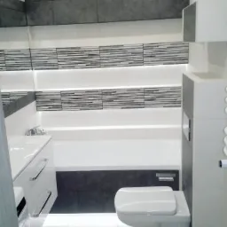 Nowoczesna łazienka z szarymi i białymi płytkami, podświetlanymi półkami, wiszącą toaletą i umywalką w zabudowie.