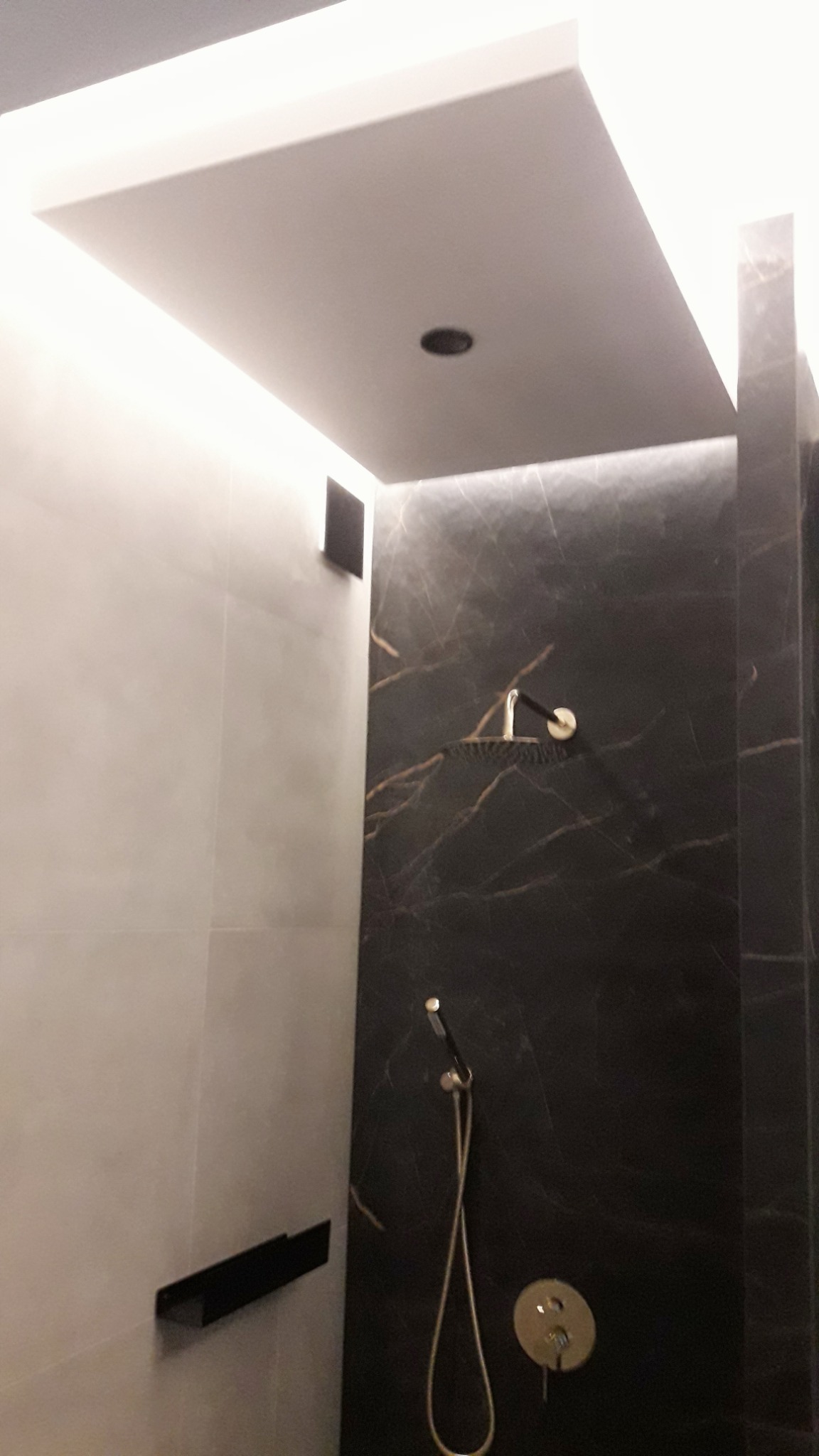 Nowoczesna kabina prysznicowa z czarną marmurową ścianą, deszczownicą i półką na kosmetyki, oświetlenie LED w suficie podwieszanym.