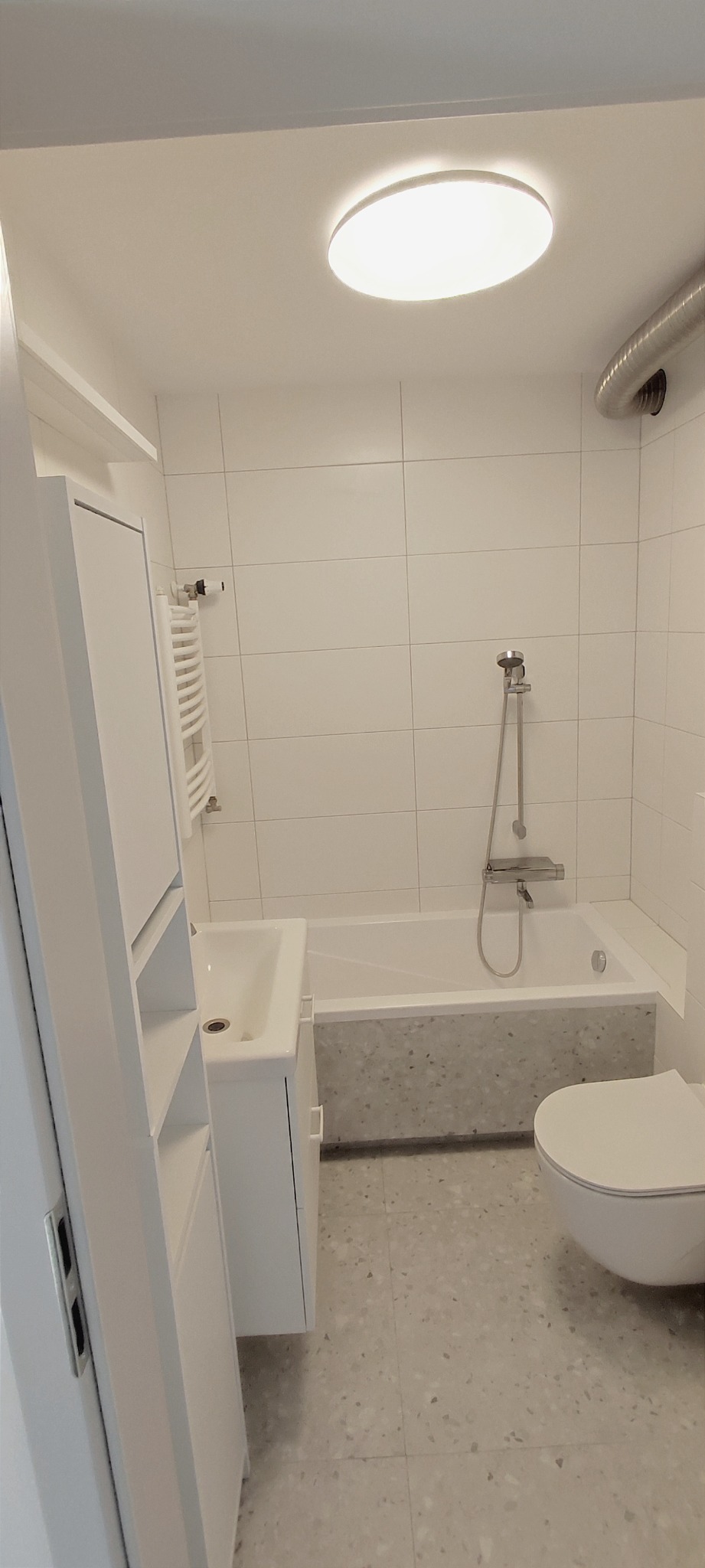 Jasna, minimalistyczna łazienka z białymi płytkami, wanną obudowaną płytami imitującymi lastryko, umywalką z szafką, toaletą i grzejnikiem drabinkowym. Okrągła lampa sufitowa.