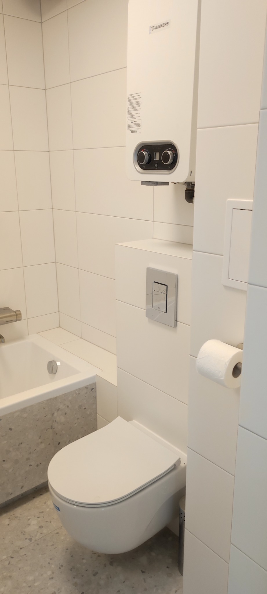 Nowoczesna, minimalistyczna łazienka z białymi płytkami, wiszącą toaletą, bojlerem Junkers i wanną z szarą obudową z lastryko.