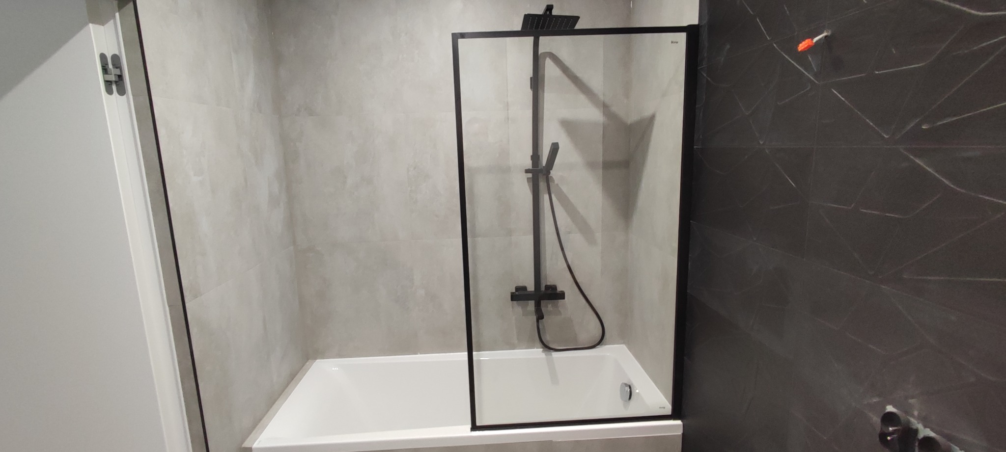 Nowoczesna łazienka z białą wanną, czarną szklaną kabiną prysznicową i szarą płytką imitującą beton na ścianach, z widocznymi elementami instalacji na ciemniejszej ścianie.