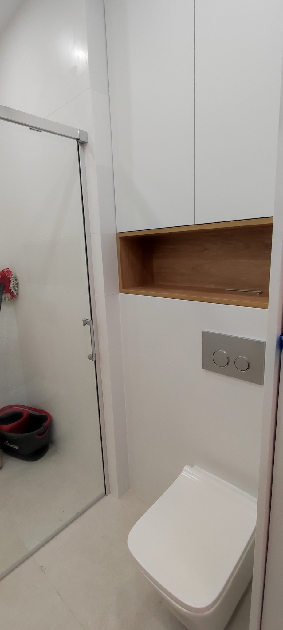 Nowoczesna toaleta z białymi płytkami, zabudową meblową z drewnianą wnęką i chromowanym przyciskiem spłukiwania. W tle widoczna szklana kabina prysznicowa z wiadrem i mopem.