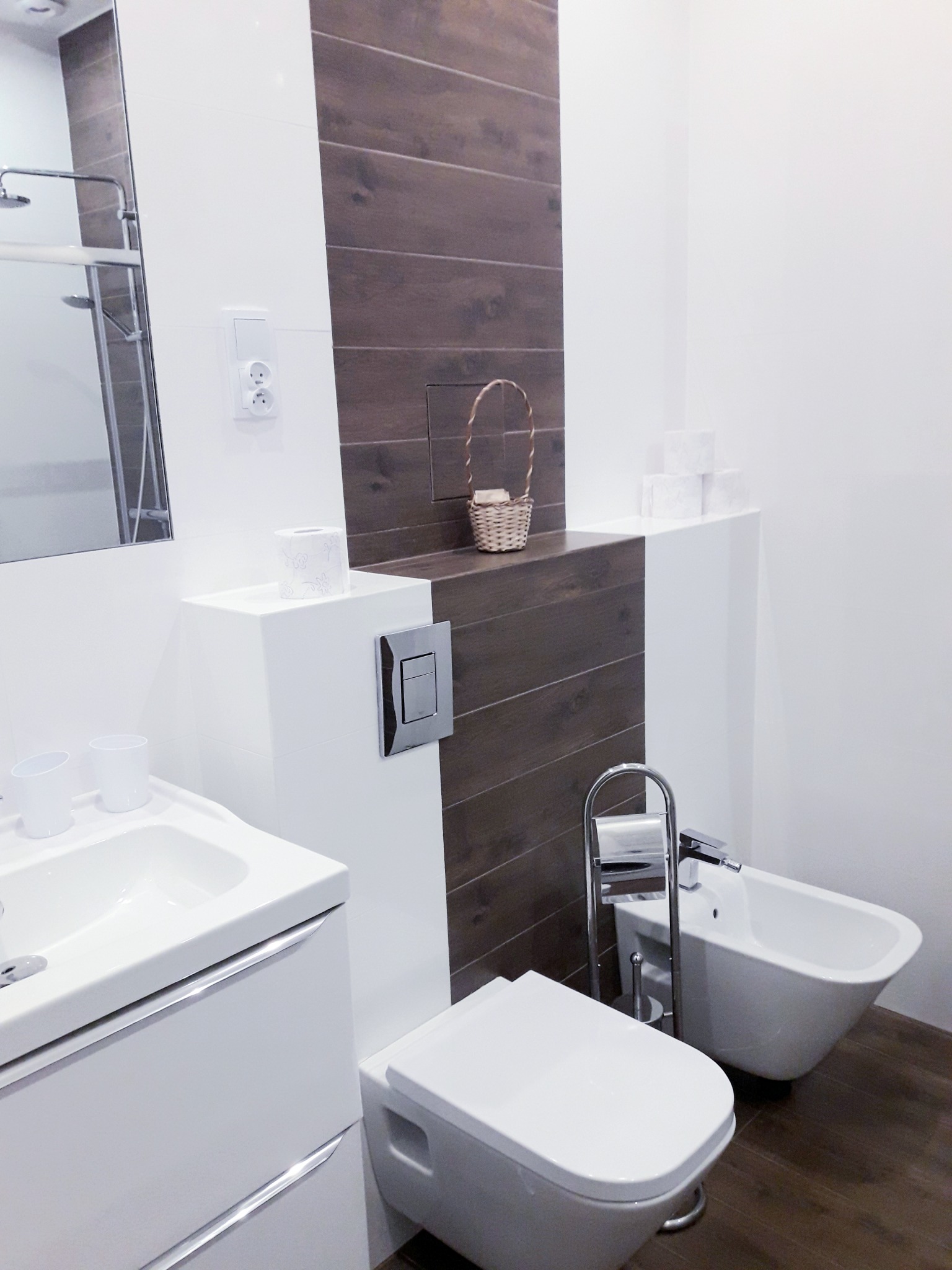 Nowoczesna łazienka w bieli z akcentem drewnopodobnych płytek, obejmująca umywalkę z szafką, toaletę, bidet i koszyk wiklinowy na półce.