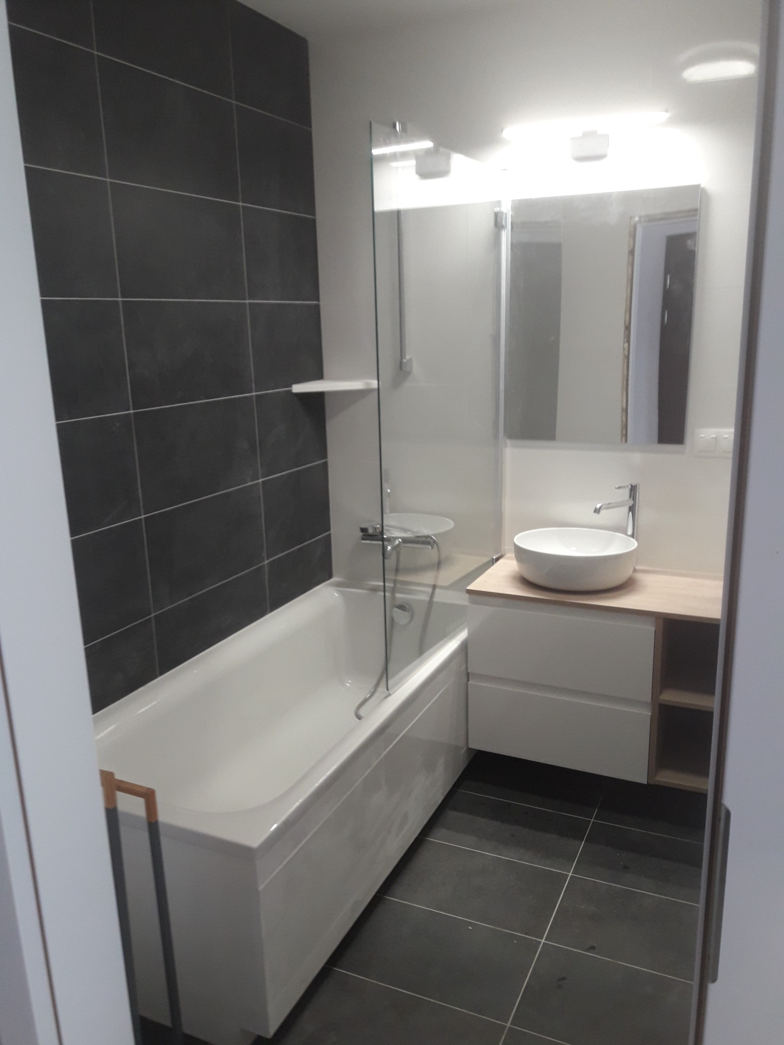 Nowoczesna łazienka z wanną obudowaną płytkami, umywalką nablatową na szafce z jasnego drewna, dużym lustrem i szarą płytką na ścianie.