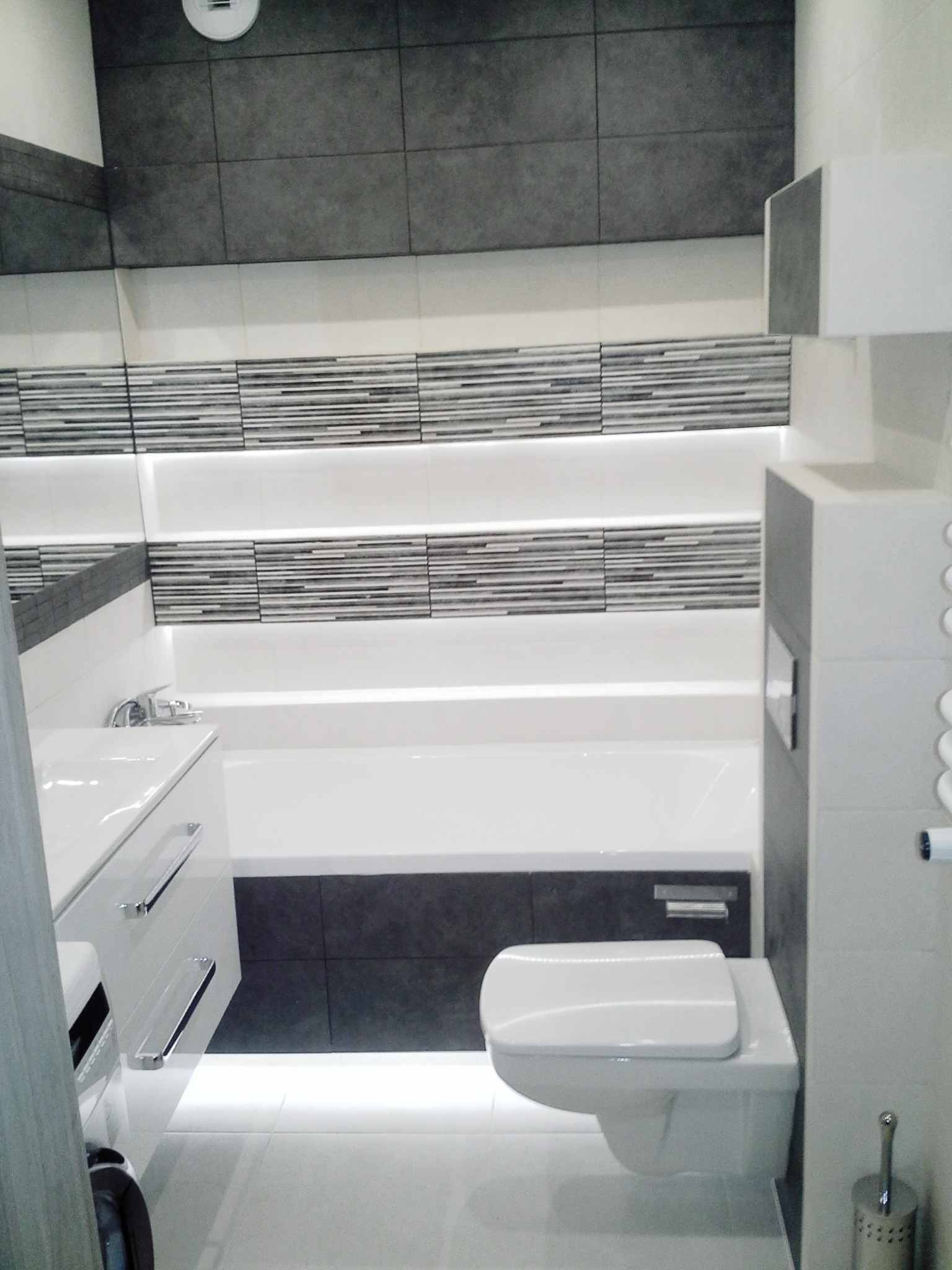 Nowoczesna łazienka z szarymi i białymi płytkami, podświetlanymi półkami, wiszącą toaletą i umywalką w zabudowie.