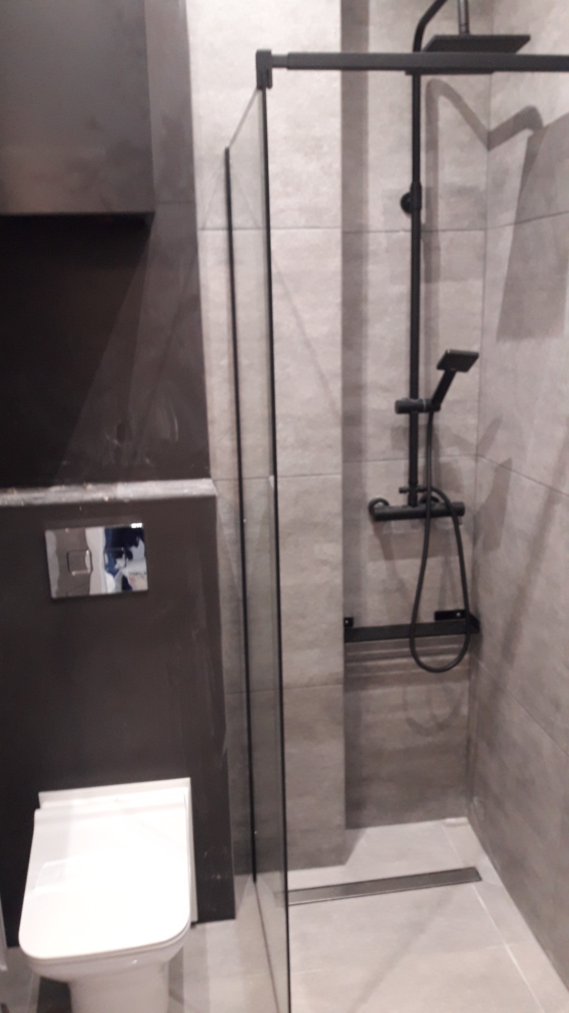 Nowoczesna łazienka z kabiną prysznicową typu walk-in z czarną armaturą, podwieszaną toaletą i szarą glazurą.