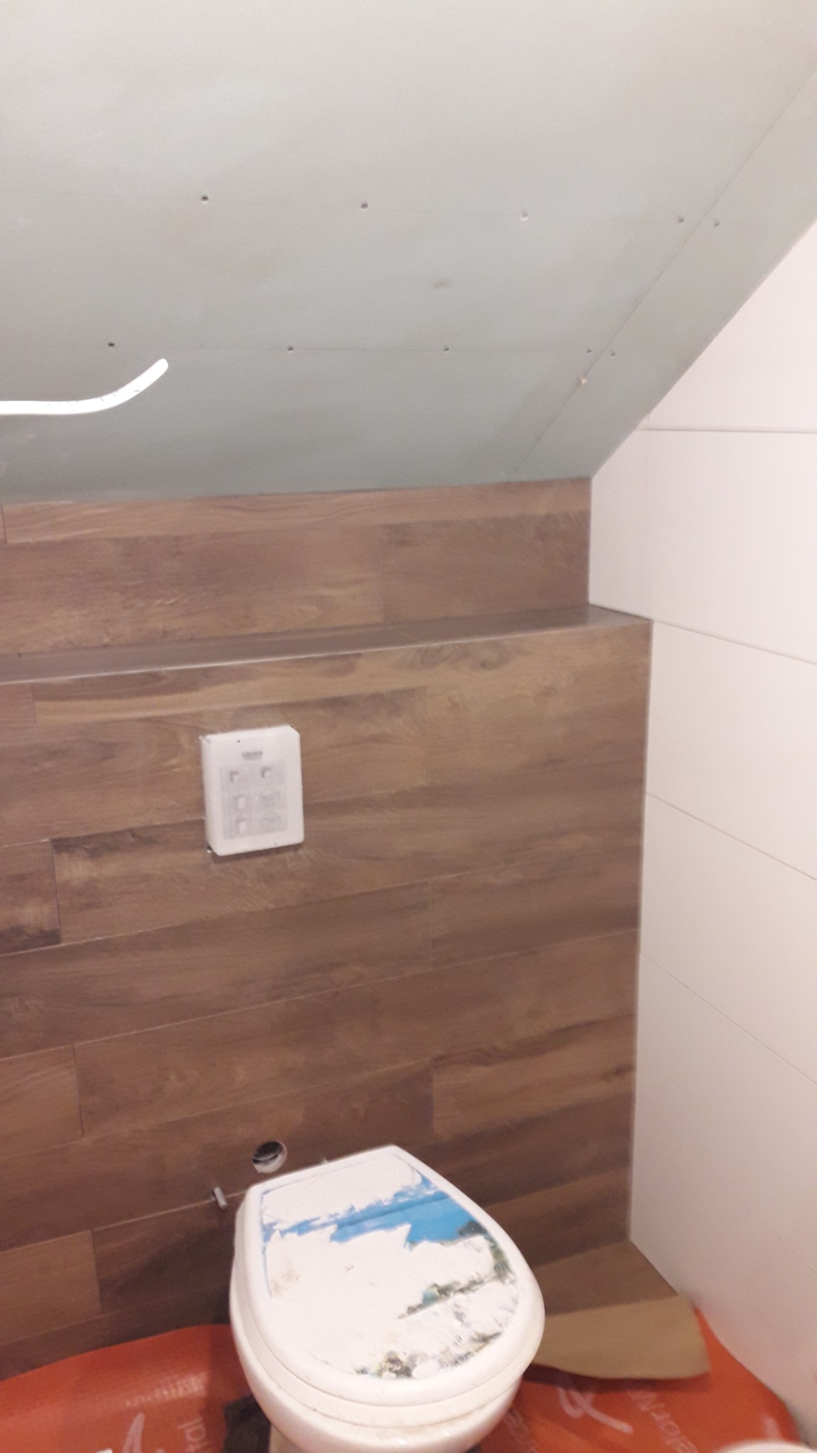 Fragment łazienki w trakcie prac wykończeniowych: brązowe płytki imitujące drewno na ścianie, częściowo zamontowana toaleta, biały panel sterujący spłuczką podtynkową i surowy sufit z płyt...