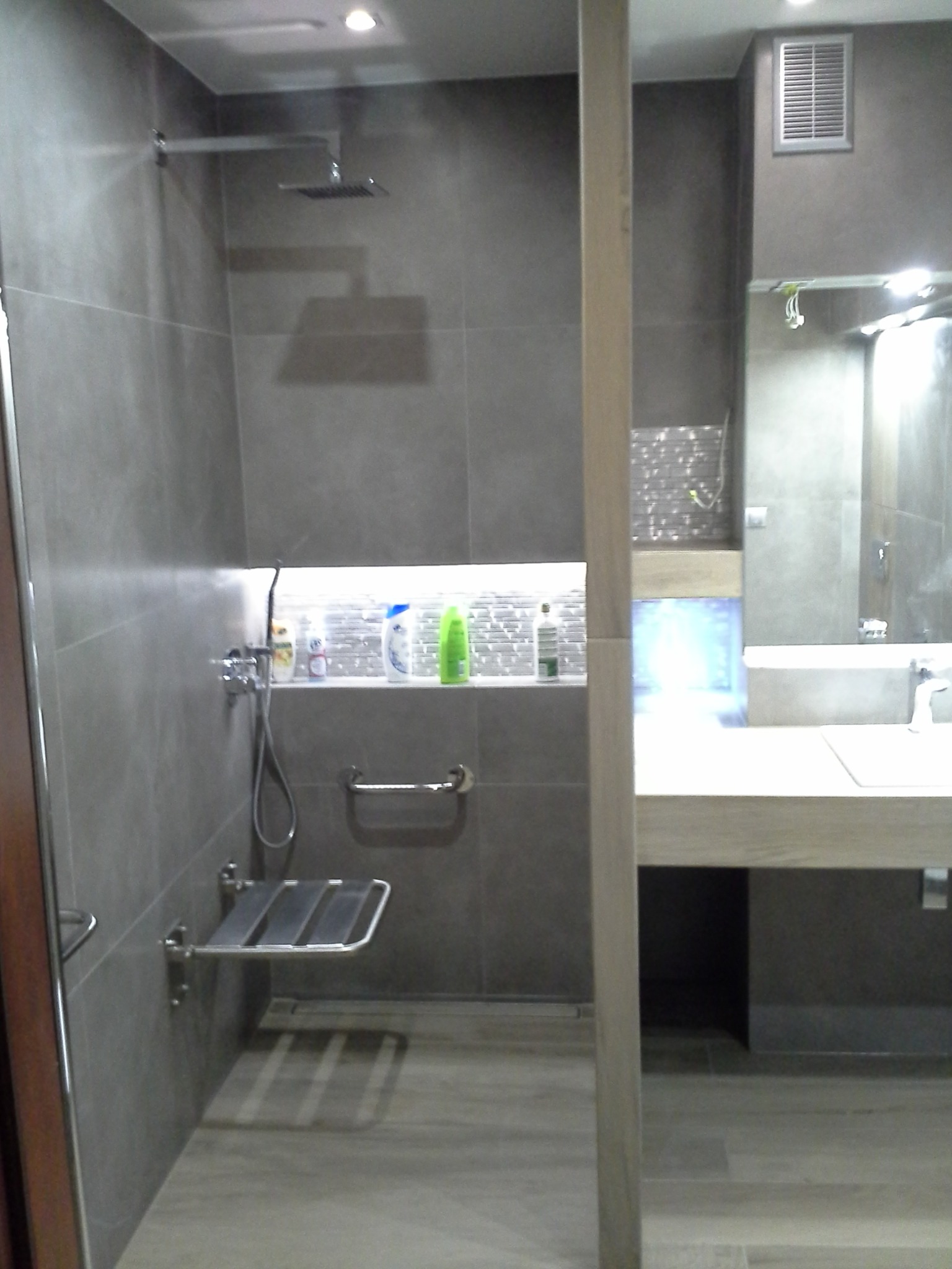 Szara łazienka z kabiną prysznicową typu walk-in, wyposażona w składane siedzisko i uchwyt dla osób z ograniczoną mobilnością, oraz półkę z kosmetykami podświetloną od dołu.