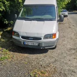 Biały Ford Transit z widocznym wgnieceniem na masce, zaparkowany na poboczu obok drzew, z polską tablicą rejestracyjną PO 118UC.
