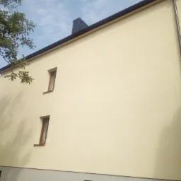 Dwupiętrowy budynek z jasną elewacją o delikatnej fakturze, z dwoma małymi oknami na górnej kondygnacji i szarym pasem u dołu, widoczny fragment dachu, komin i drzewa.