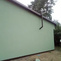 Jasnozielona elewacja budynku jednorodzinnego z brązowym orynnowaniem i kominem wentylacyjnym, widoczny mężczyzna w tle, puste butelki i wiadro na ziemi.