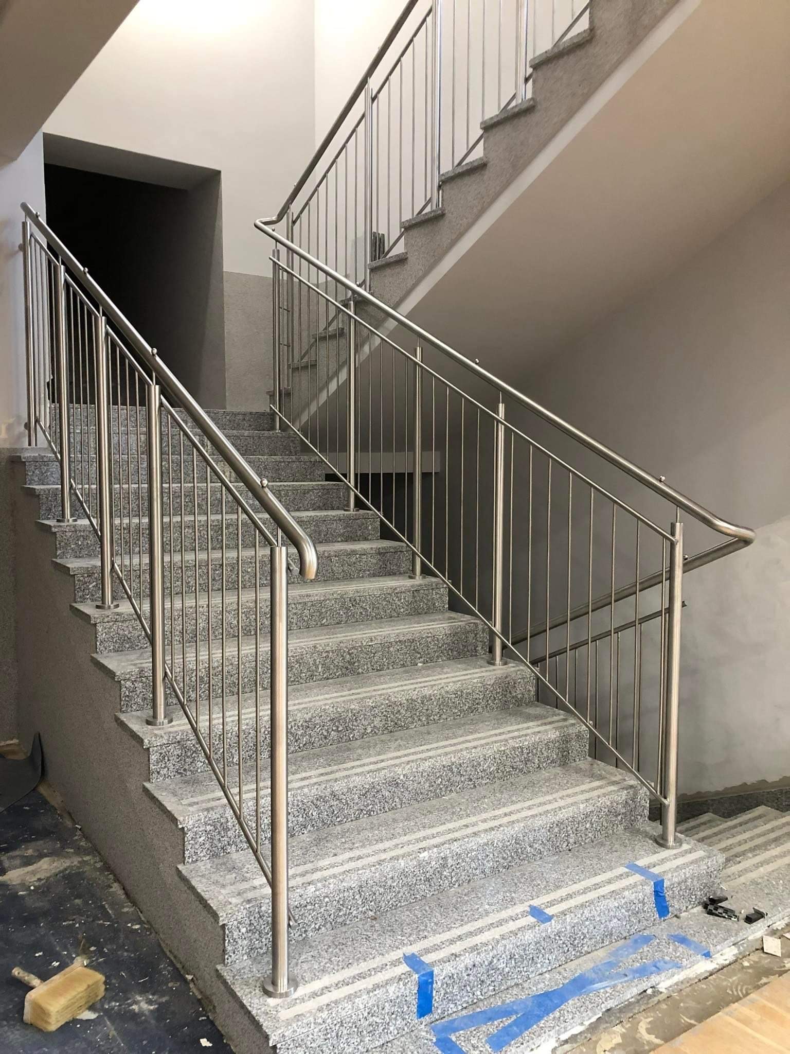 Wewnętrzna klatka schodowa z szarymi, lastrykowymi schodami i metalową poręczą, widoczne ślady prac wykończeniowych, zabezpieczenia taśmą malarską.