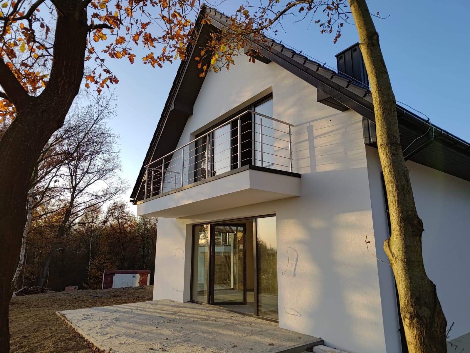 Nowoczesny dom z balkonem zabezpieczonym metalową konstrukcją z cienkich prętów, widok od strony tarasu z otwartymi drzwiami balkonowymi, jesienne drzewa w tle.