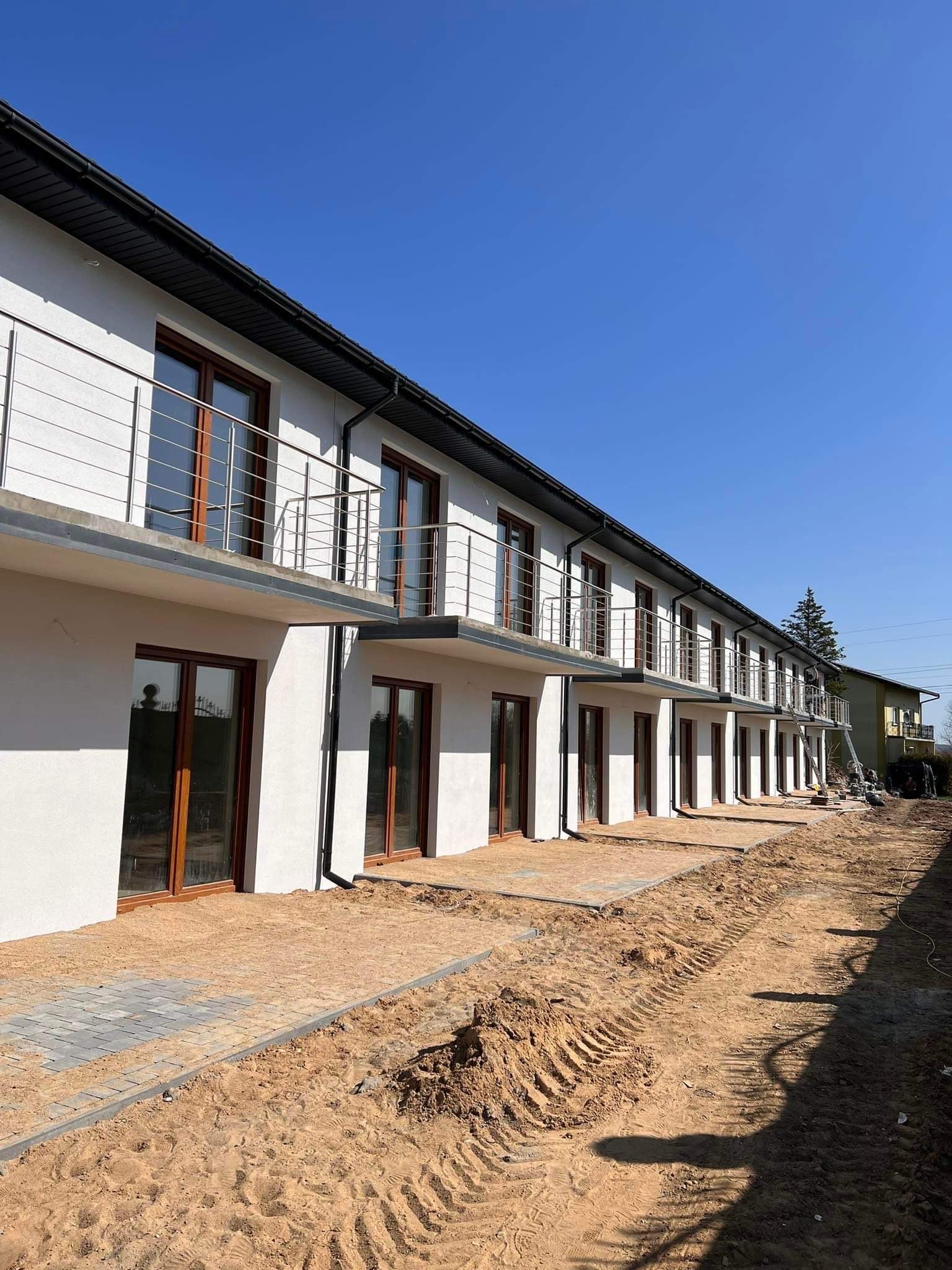 Nowe budynki mieszkalne z balkonami zabezpieczonymi prostymi, metalowymi barierkami, widok z poziomu gruntu na placu budowy z piaskiem i częściowo ułożoną kostką.