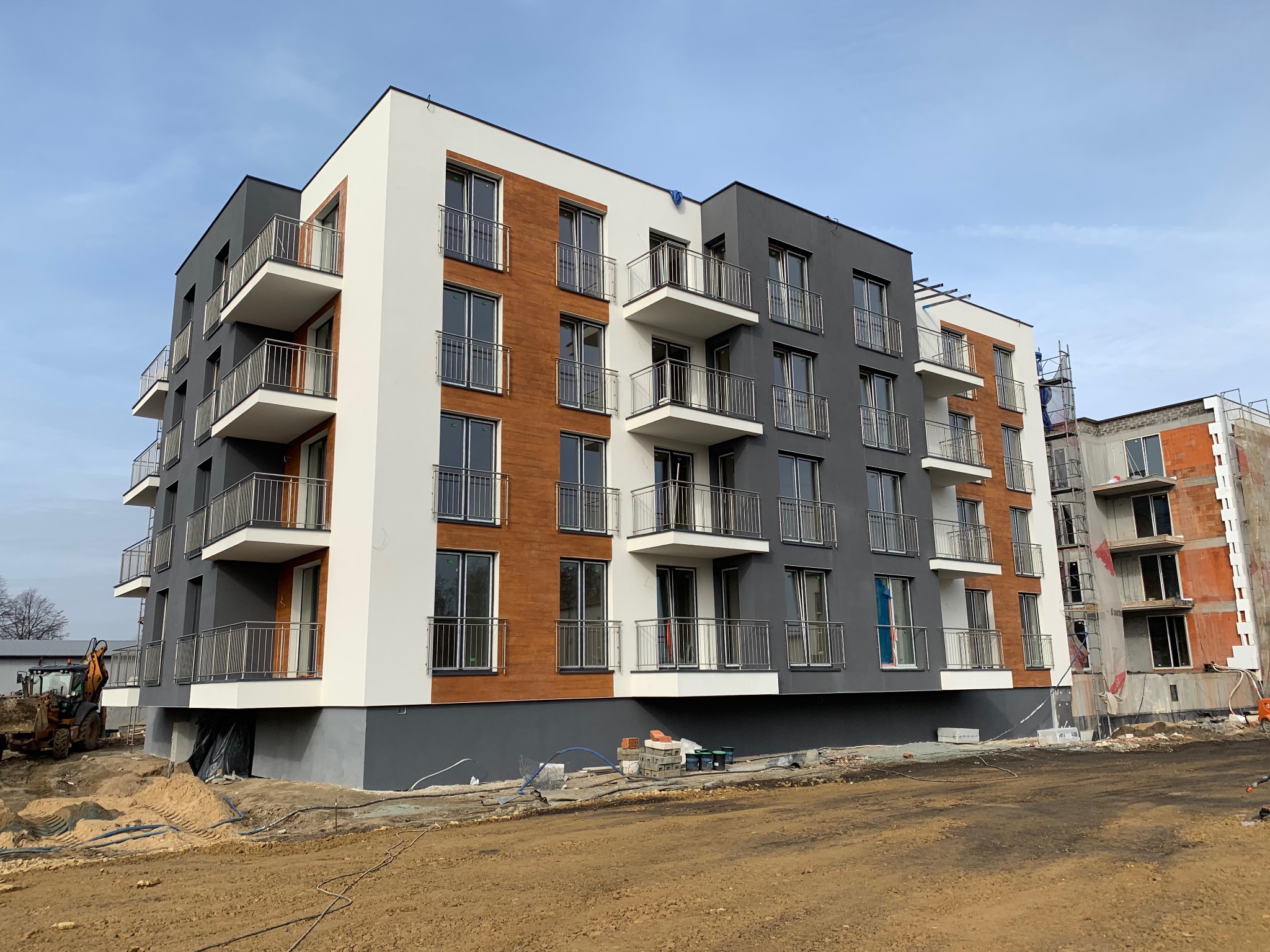 Nowoczesny budynek wielorodzinny z balkonami zabezpieczonymi metalowymi barierkami, widok z poziomu gruntu na placu budowy.