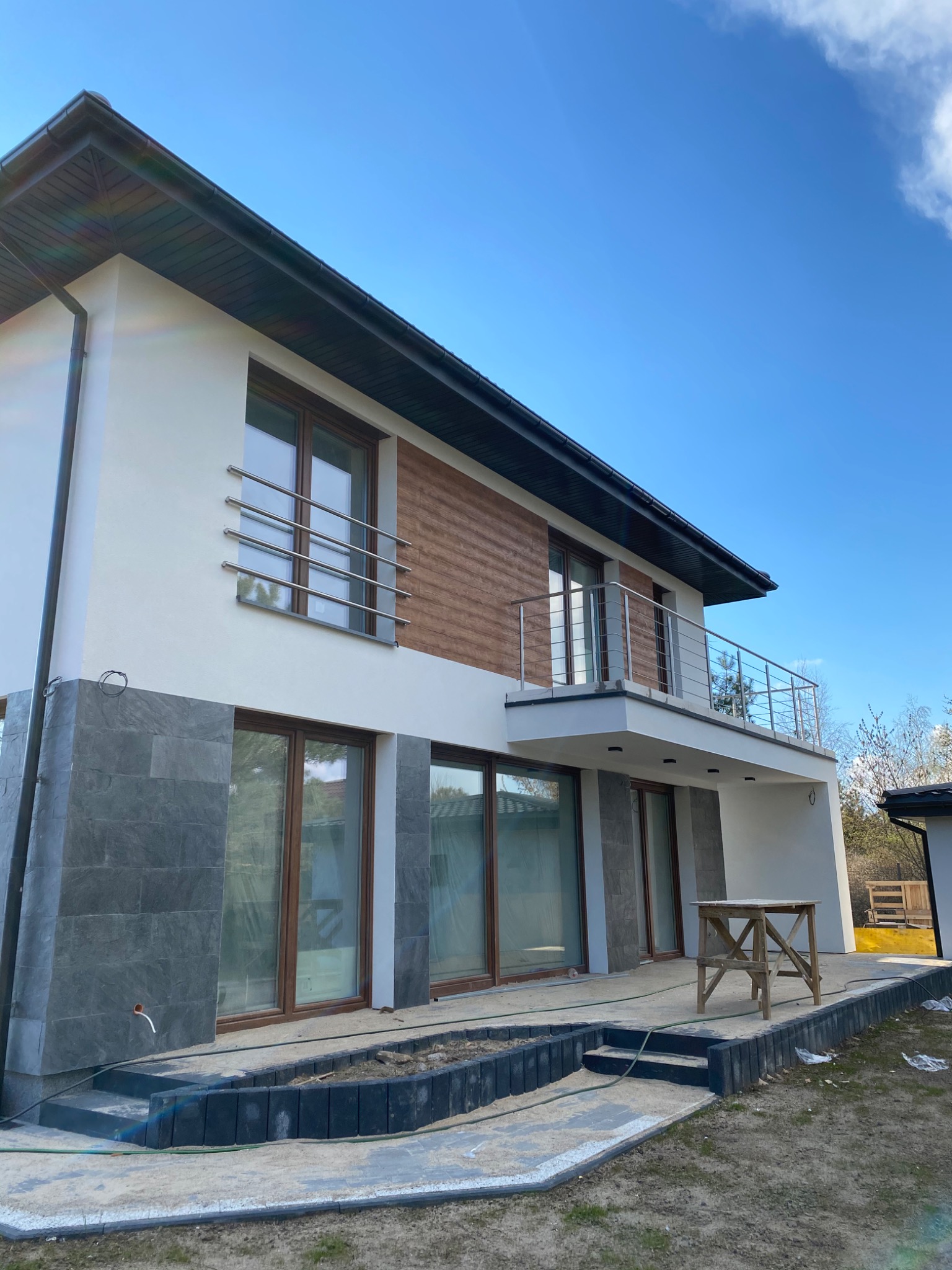 Nowoczesny dom z balkonem z balustradą nierdzewną, widok z dołu na elewację z elementami drewna i kamienia, teren wokół domu w trakcie prac wykończeniowych, widoczne rusztowanie.