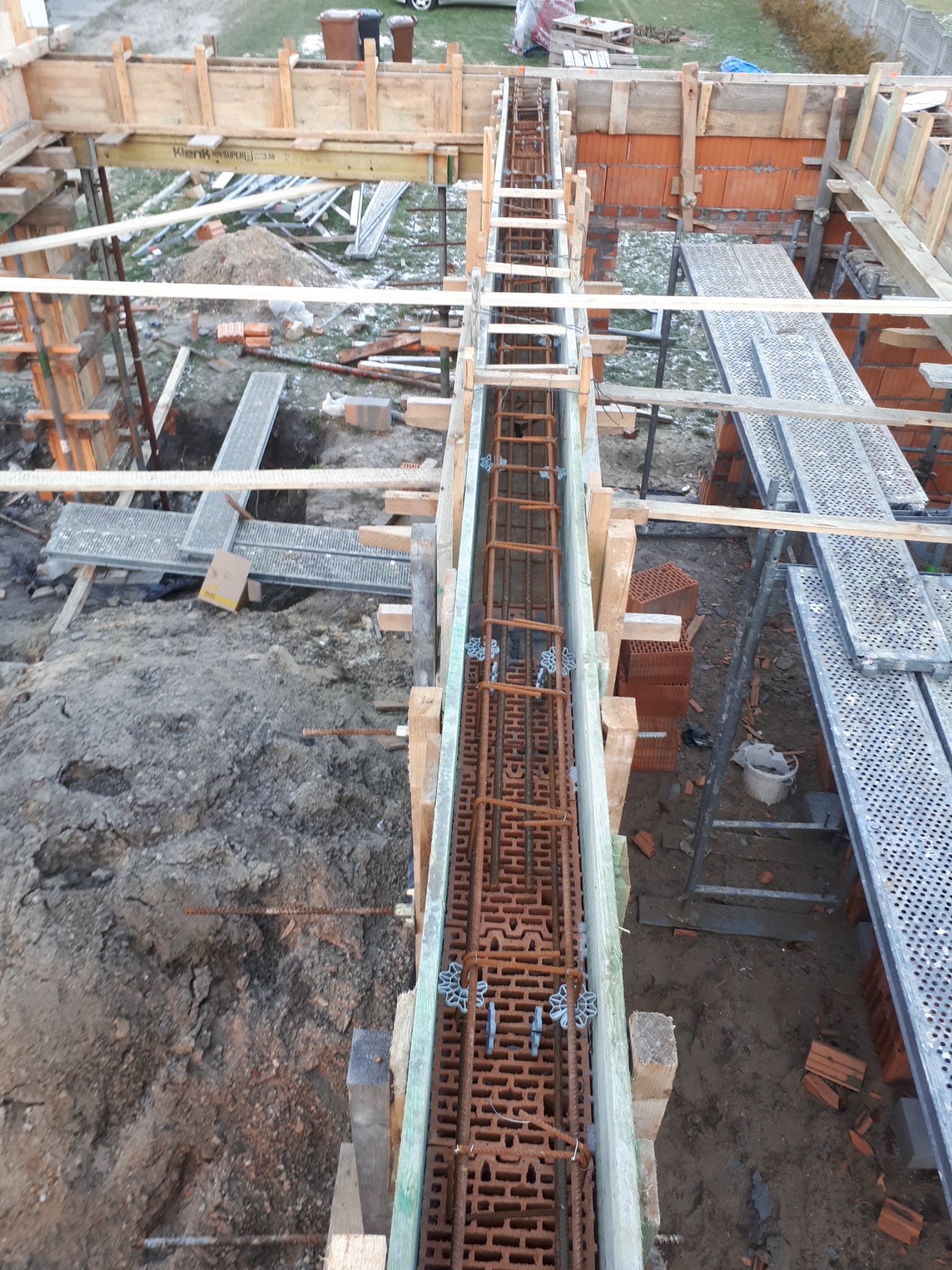 Zbrojenie w szalunku pod wylewkę fundamentu, widoczne metalowe rozpórki i cegły dziurawki wewnątrz konstrukcji, rusztowanie w tle.