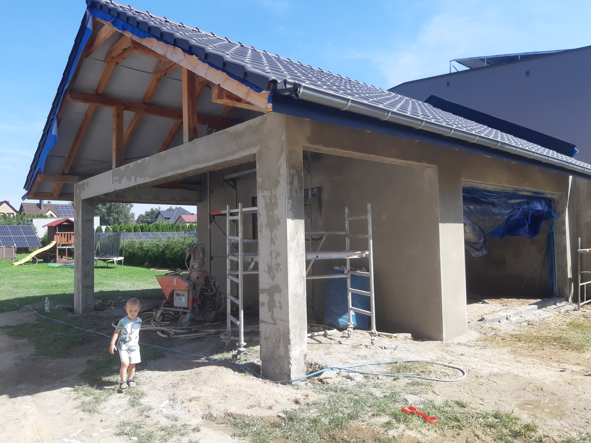 Budowa garażu z betonowych bloczków i dachem z dachówki ceramicznej. Na pierwszym planie dziecko, w tle plac zabaw i domy.