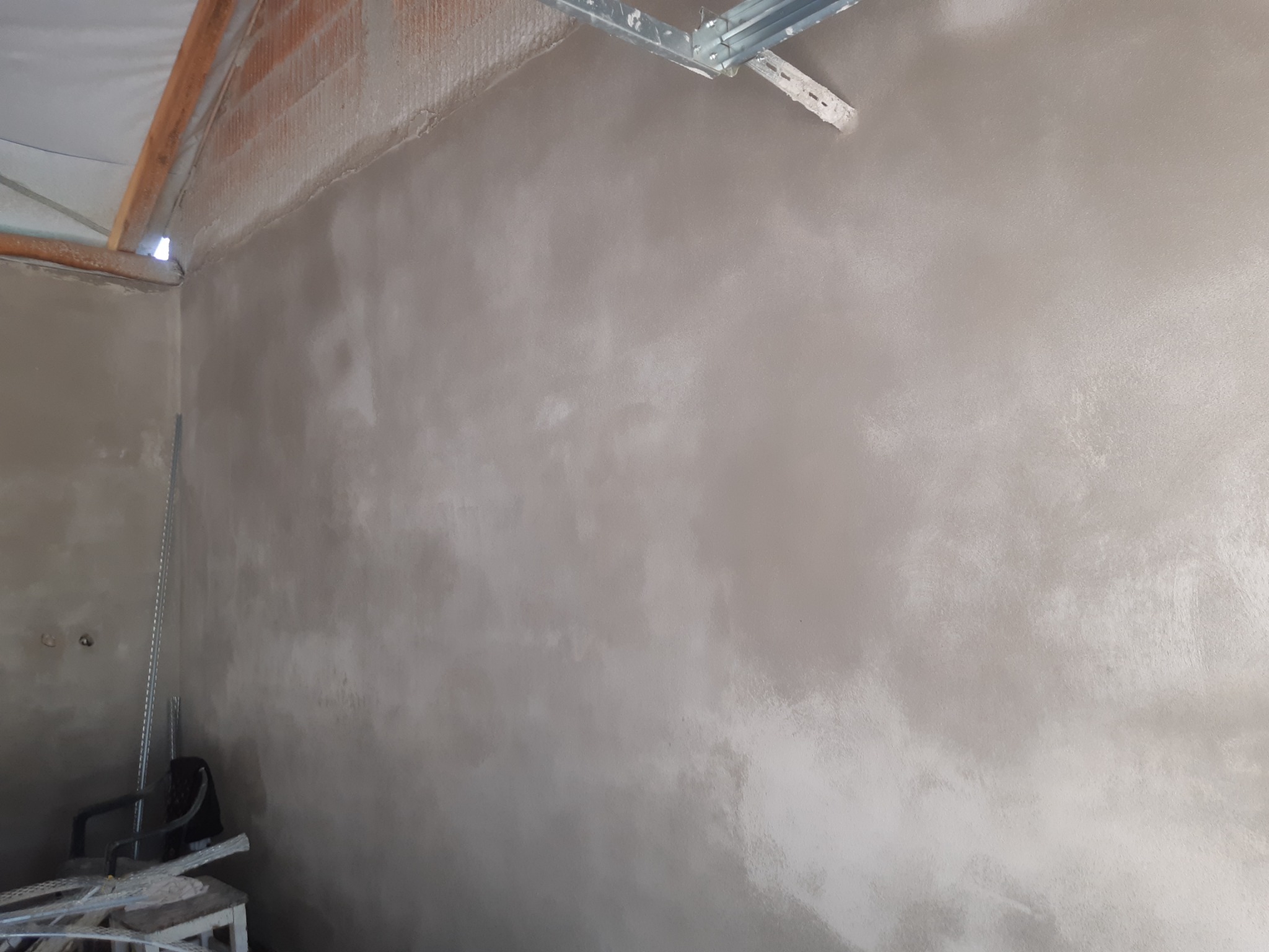 Świeżo otynkowana ściana wewnętrzna z widocznymi elementami konstrukcyjnymi sufitu i narzędziami w rogu pomieszczenia.