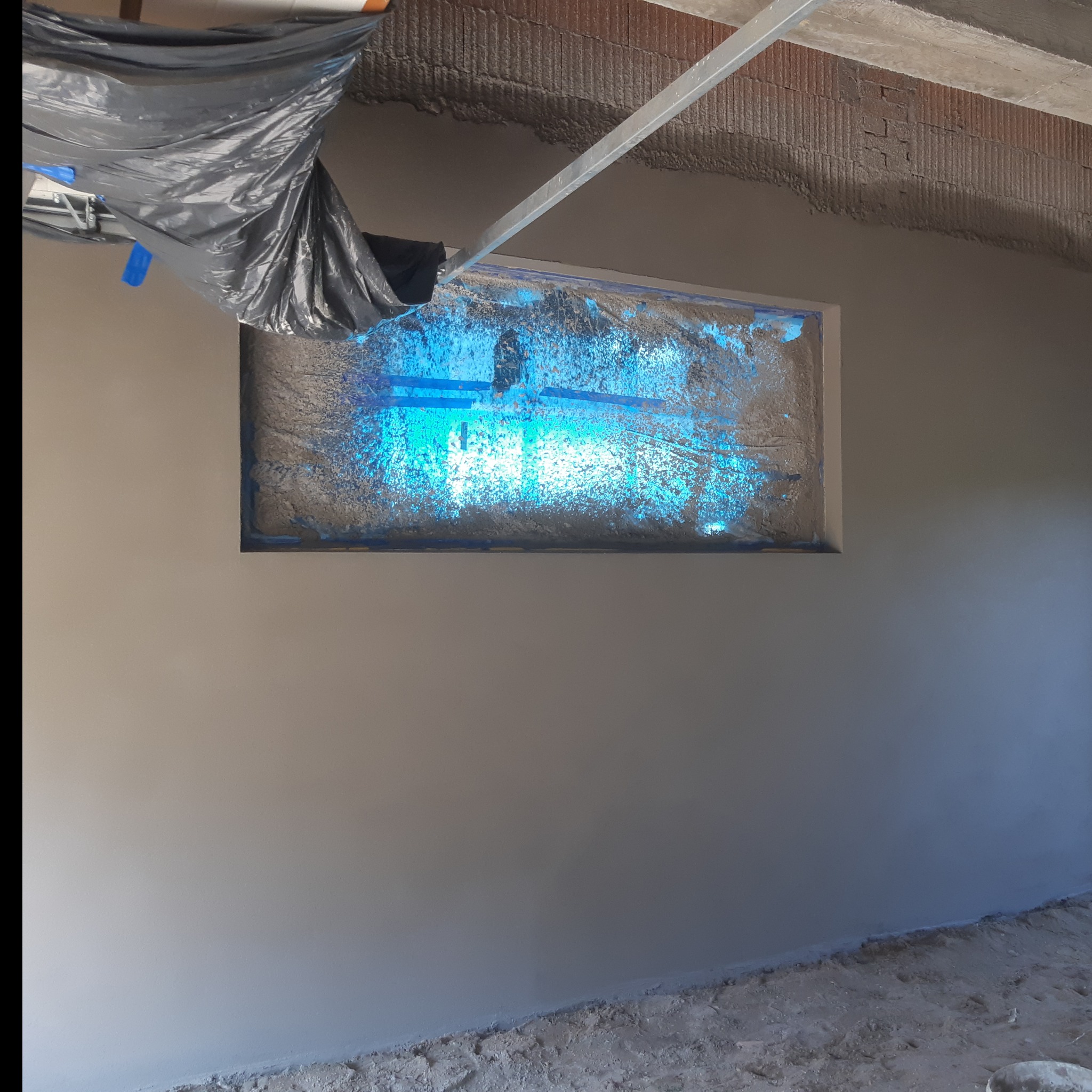 Świeżo otynkowana ściana wewnętrzna z zabezpieczonym oknem folią i resztkami zaprawy, widoczny surowy strop z instalacją.