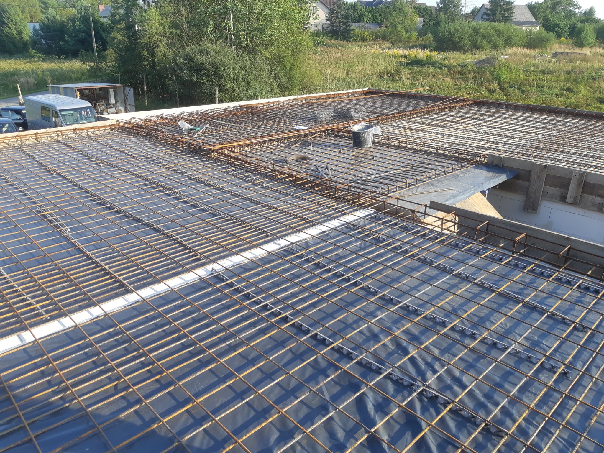 Siatka zbrojeniowa na folii izolacyjnej, przygotowana do wylania fundamentów, widok z góry, w tle zabudowa jednorodzinna i zieleń.