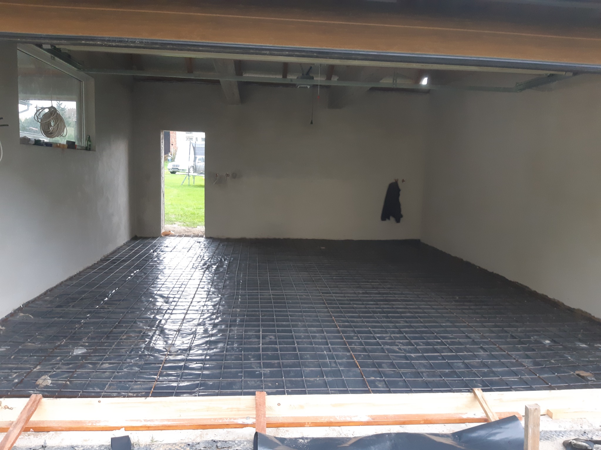 Przygotowanie podłoża pod wylewkę: siatka zbrojeniowa na czarnej folii w surowym wnętrzu budynku z widocznymi elementami konstrukcyjnymi i otwartym przejściem na zewnątrz.