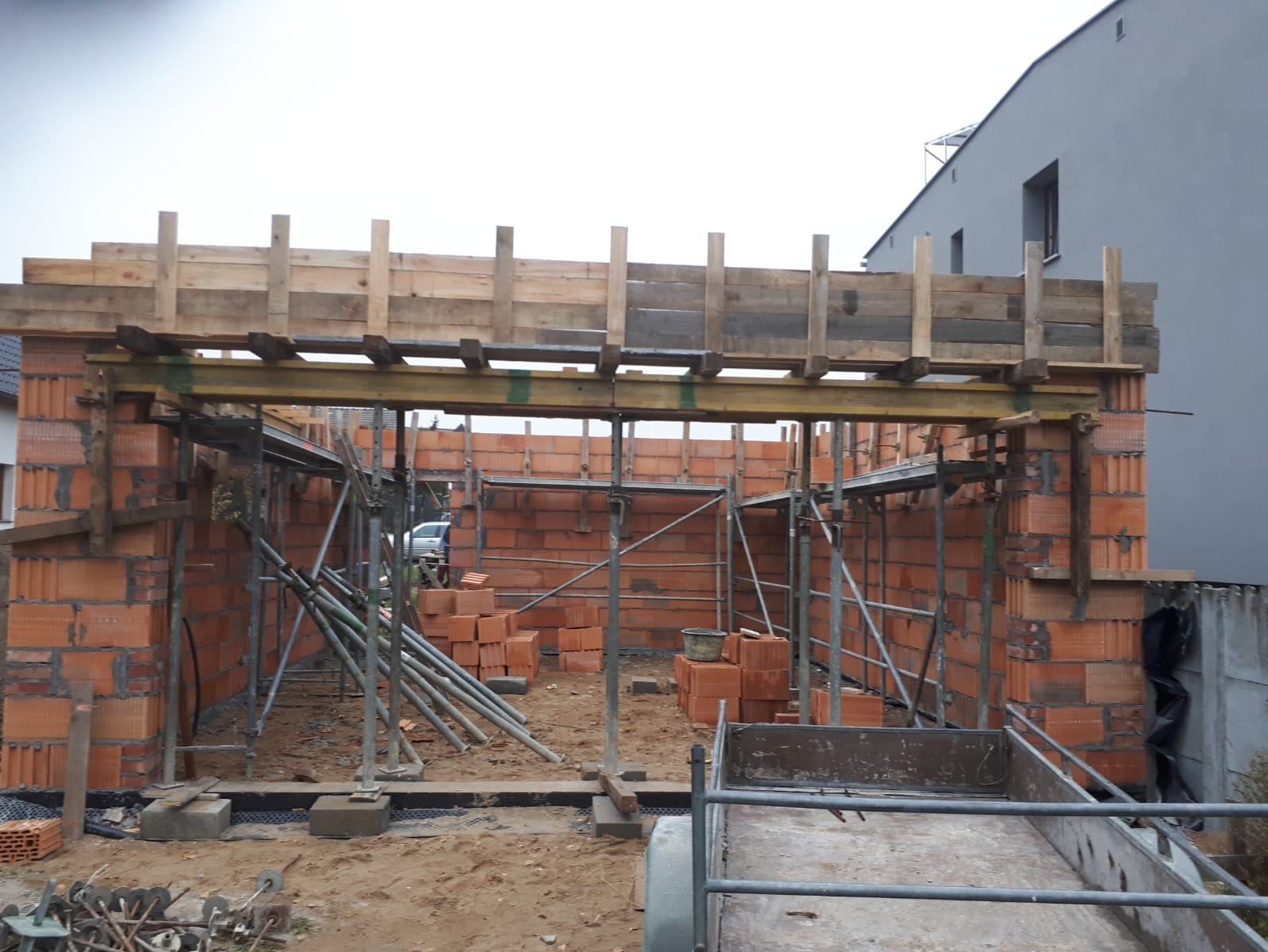 Wznoszenie ścian z czerwonej cegły na budowie domu jednorodzinnego, widoczne rusztowania, drewniane szalunki stropu i przyczepka transportowa w tle.