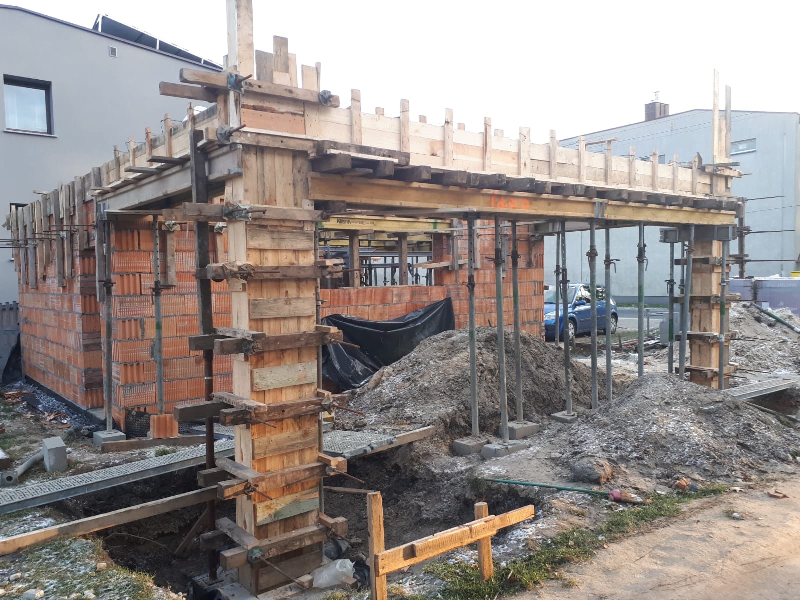 Wznoszenie murów z czerwonej cegły z drewnianym szalunkiem i metalowymi podporami, widoczny wykop i hałda ziemi, w tle budynki mieszkalne i niebieski samochód.