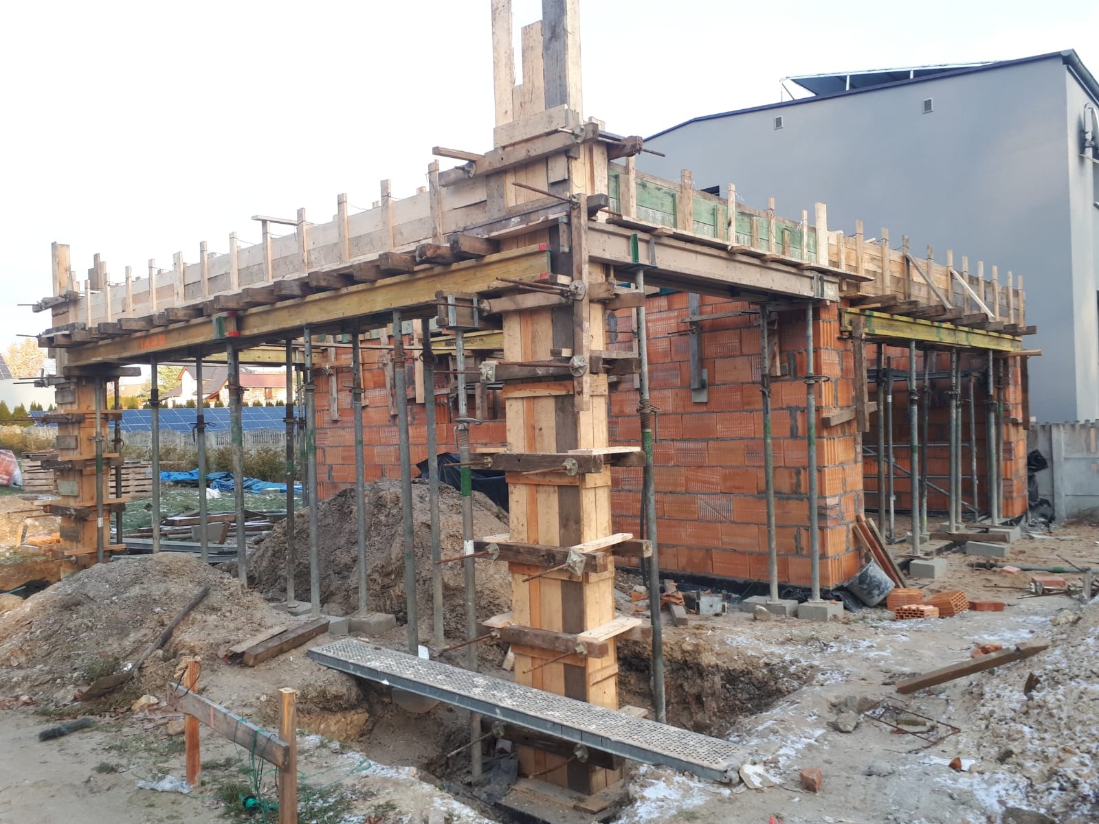 Wznoszenie ścian z czerwonej cegły z użyciem drewnianego szalunku i metalowych podpór, widoczne hałdy ziemi i sąsiedni budynek.