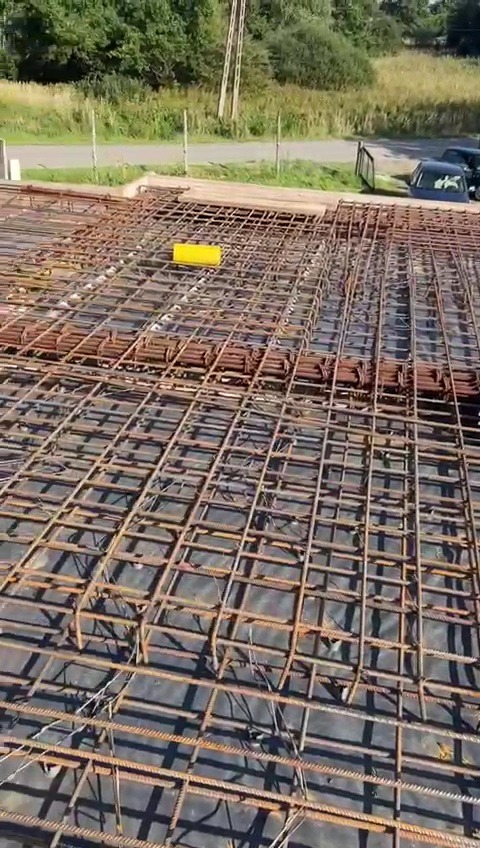 Siatka zbrojeniowa na czarnej folii, widok z góry, żółty element dystansowy, w tle zieleń i zaparkowane auta.