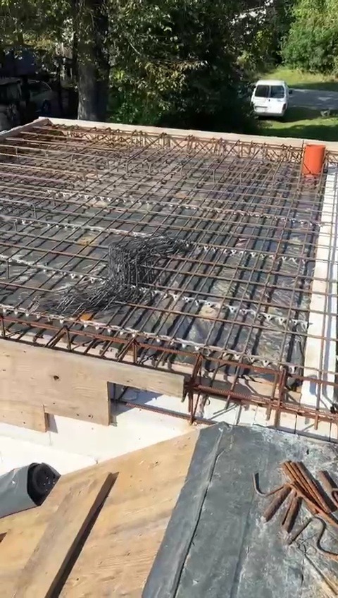 Zbrojenie stropu w trakcie budowy, widoczne pręty zbrojeniowe ułożone w kratę na folii, drewniane szalunki, w tle biały samochód dostawczy.