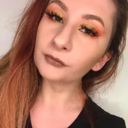 Portret kobiety z pomarańczowo-żółtym cieniowaniem powiek i małymi błyszczącymi aplikacjami przy zewnętrznym kąciku oka, brązowa szminka, włosy w odcieniach rudości.