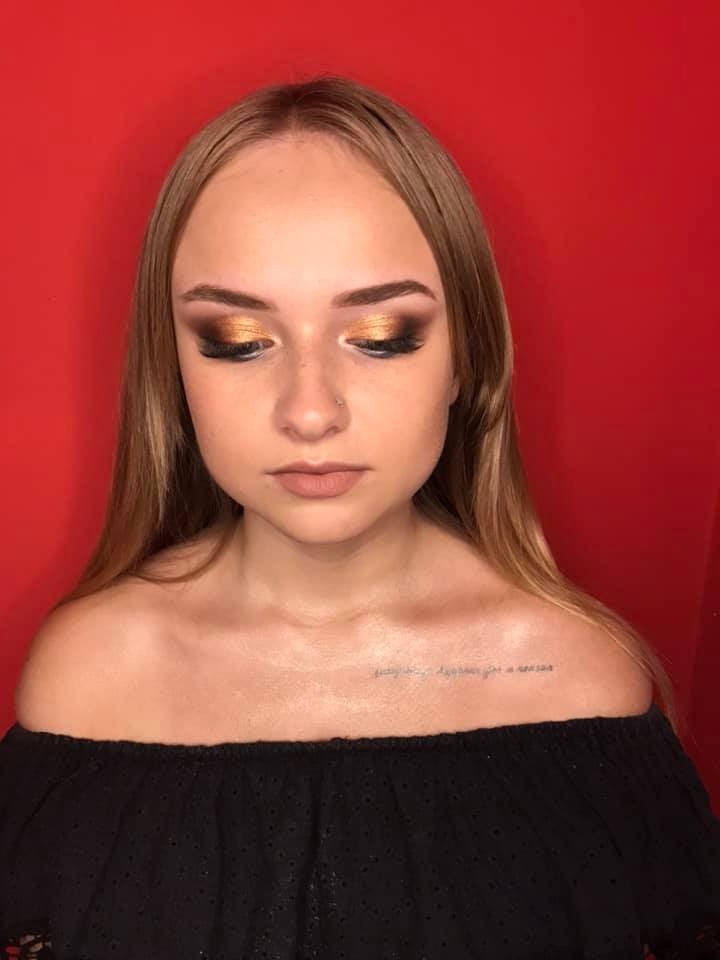 Portret kobiety z intensywnym, złotym cieniowaniem powiek i delikatnym makijażem ust, na czerwonym tle, z widocznym tatuażem na dekolcie.