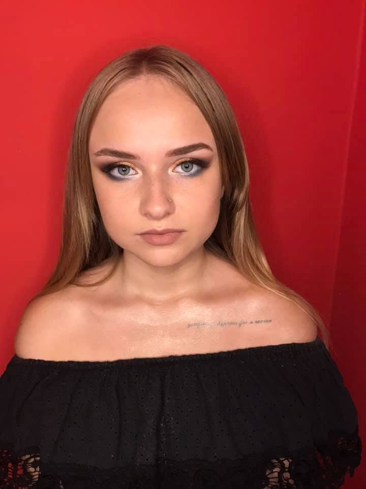 Portret kobiety z wieczorowym makijażem oczu w odcieniach szarości i złota, z delikatnym tatuażem na obojczyku, na czerwonym tle.