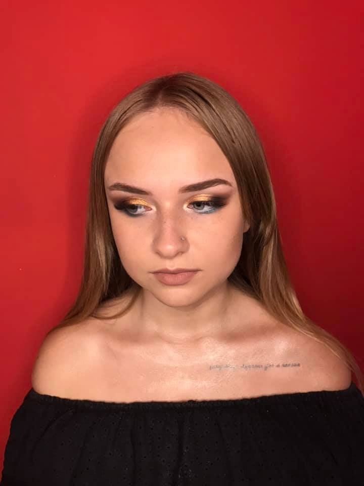 Portret kobiety z wieczorowym eye-makeup w odcieniach złota i czerni, na czerwonym tle. Widoczny delikatny tatuaż na obojczyku.