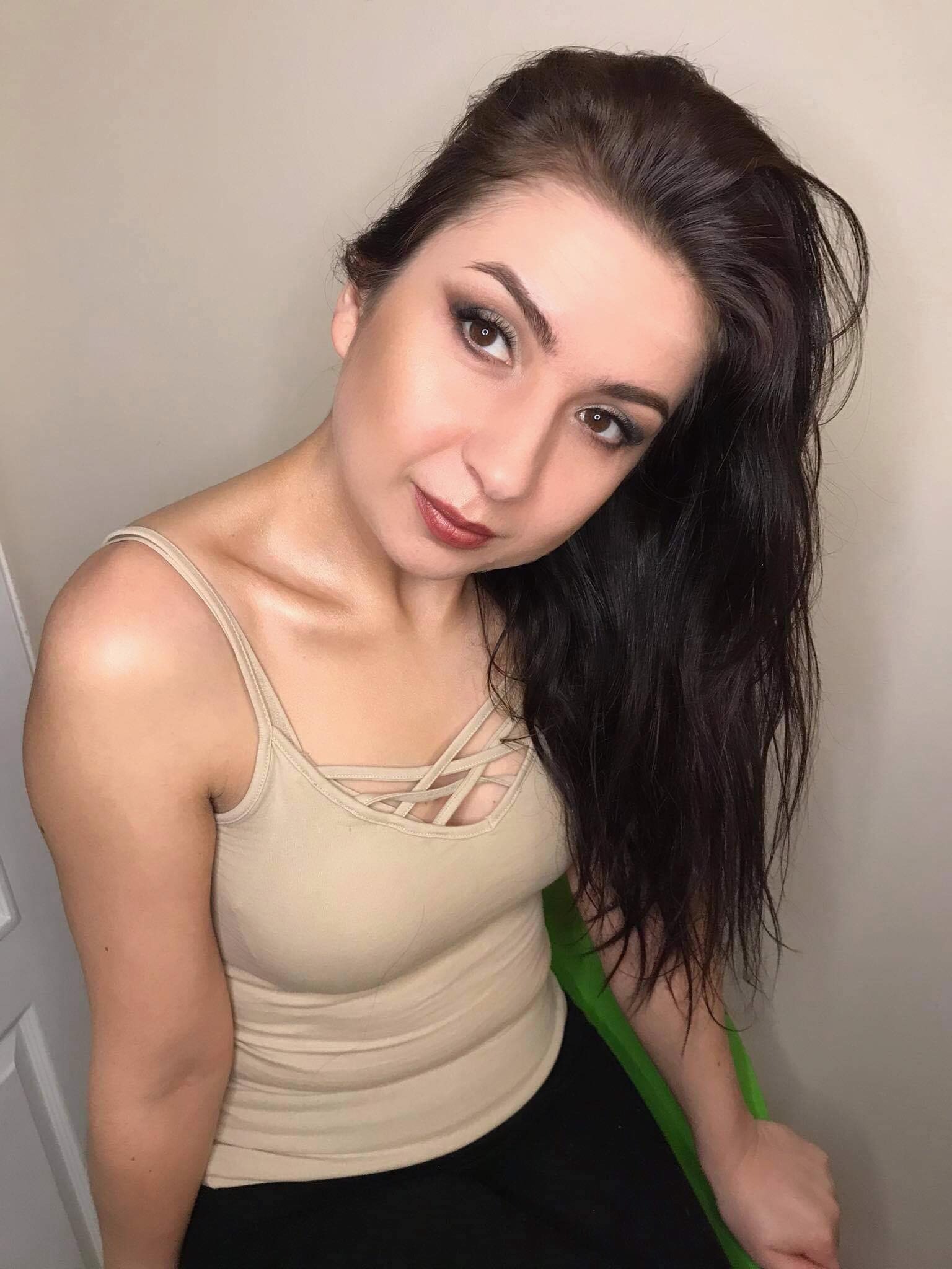 Portret kobiety z ciemnymi włosami i wyrazistym makijażem oczu, z błyszczącymi ustami w odcieniu bordo i delikatnym konturowaniem twarzy, pozująca w beżowej koszulce na tle jasnej ściany.