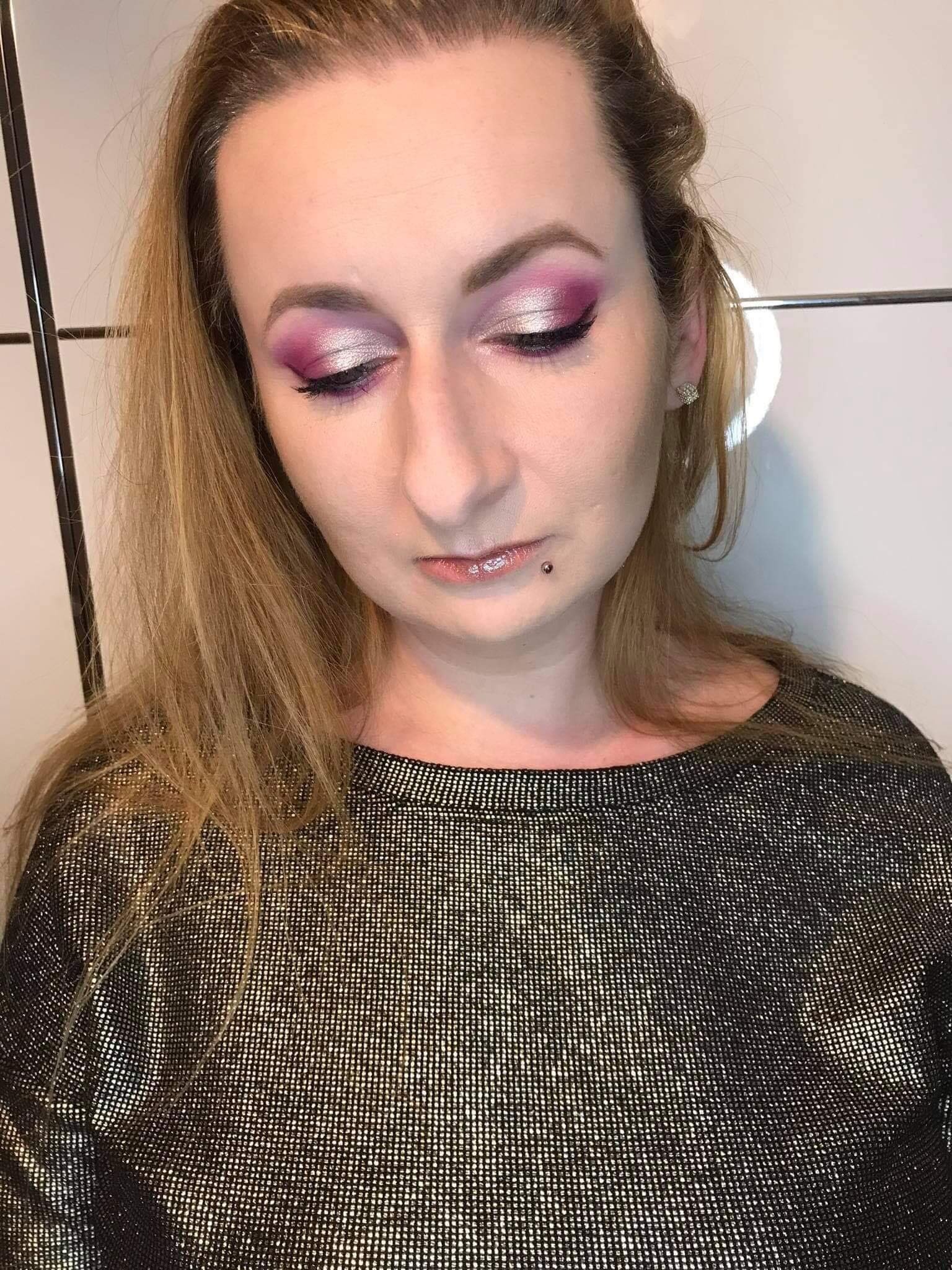 Kobieta z blond włosami i intensywnym, różowo-srebrnym cieniowaniem powiek, skierowana wzrokiem w dół, z widocznym kolczykiem w wardze i błyszczącą bluzką.